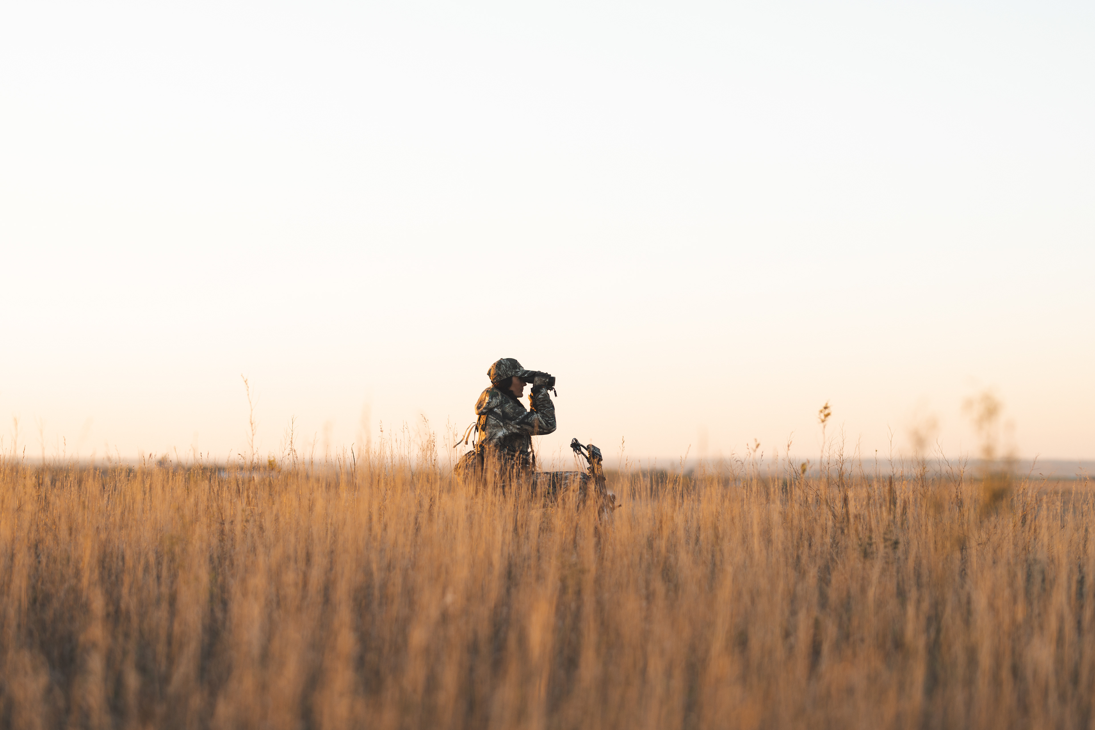 Melissa Bachman, bow hunter, in the middle of a wide shot, surveying the landscape through binoculars, in an amber field of tall brush in South Dakota