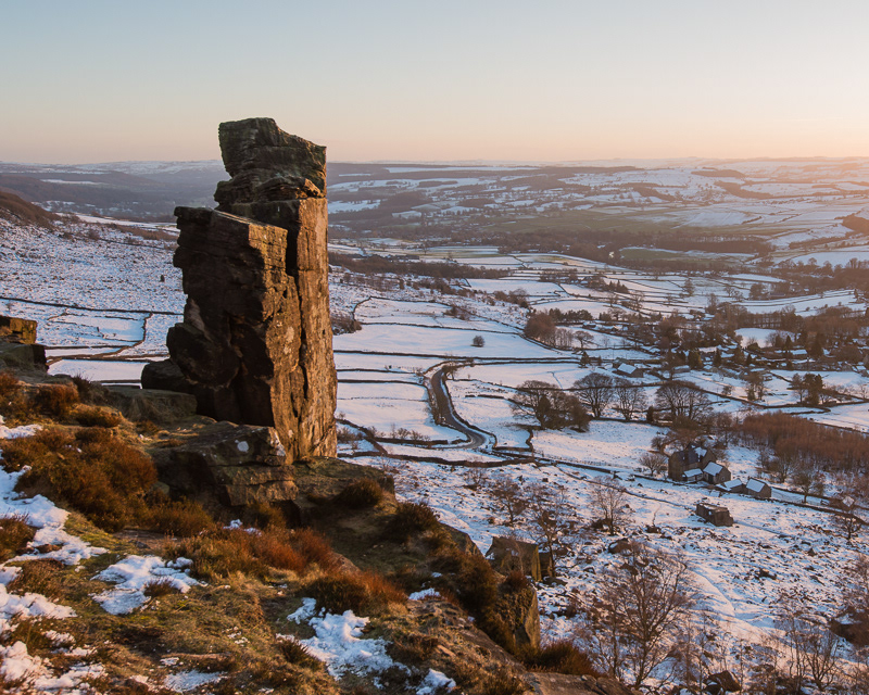 The setting sun casting warm light onto the Pinnacle on Curbar Edge above a snowy valley in the Peak District.