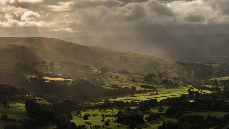 Sun rays bursting through stormy clouds casting scattered light onto the landscape of Hope Valley in the Peak District.