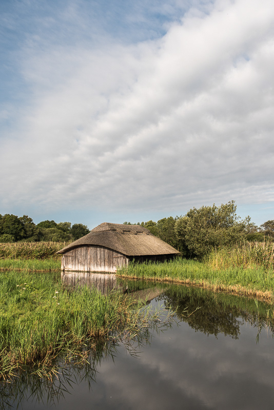 The clouds reflecting in the still water of Hickling Broad, Nrofolk with a thatched boat house in the background.