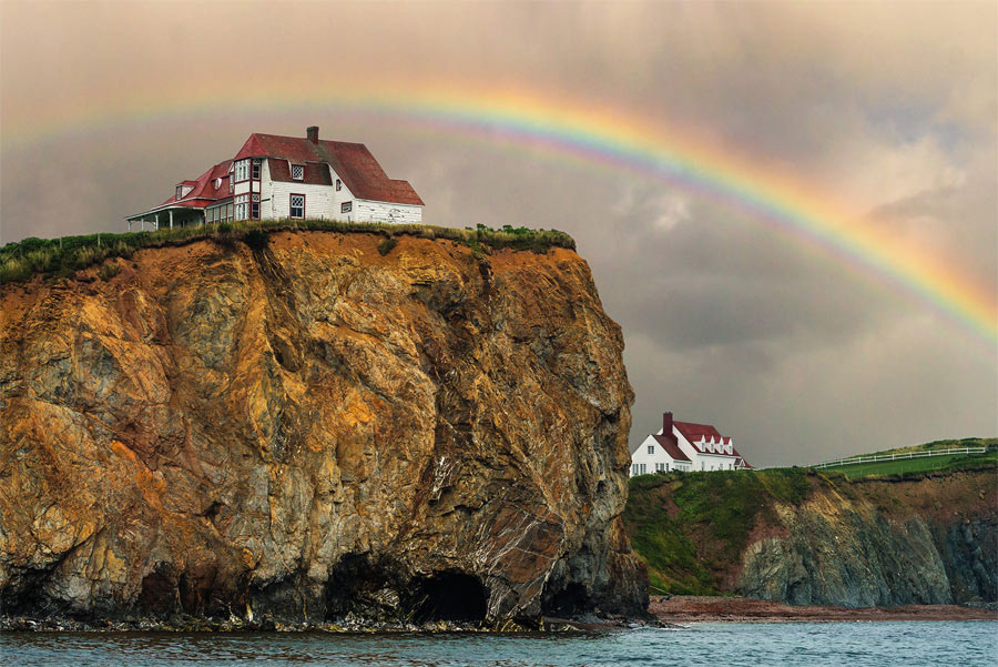 Two white houses with red roofs perched on clifftops under a rainbow, in Perce, QC