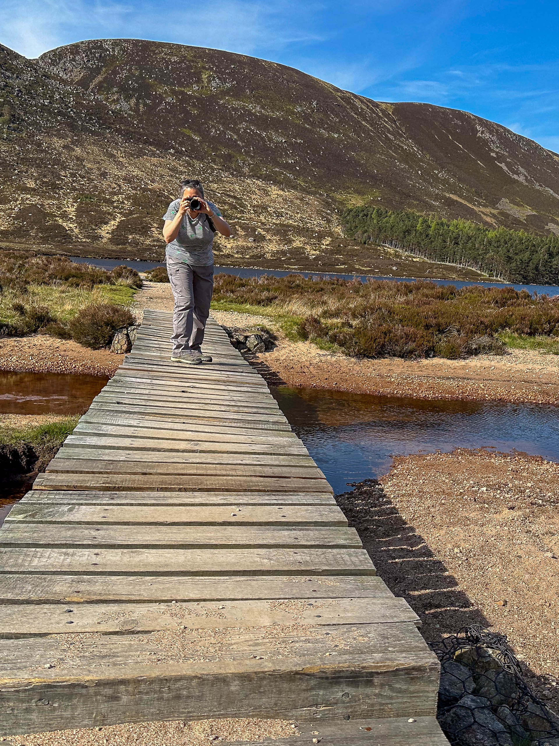 Jael taking a photo with her camera directly to the viewer at loch Muick standing on a slated bridge on a summer day.