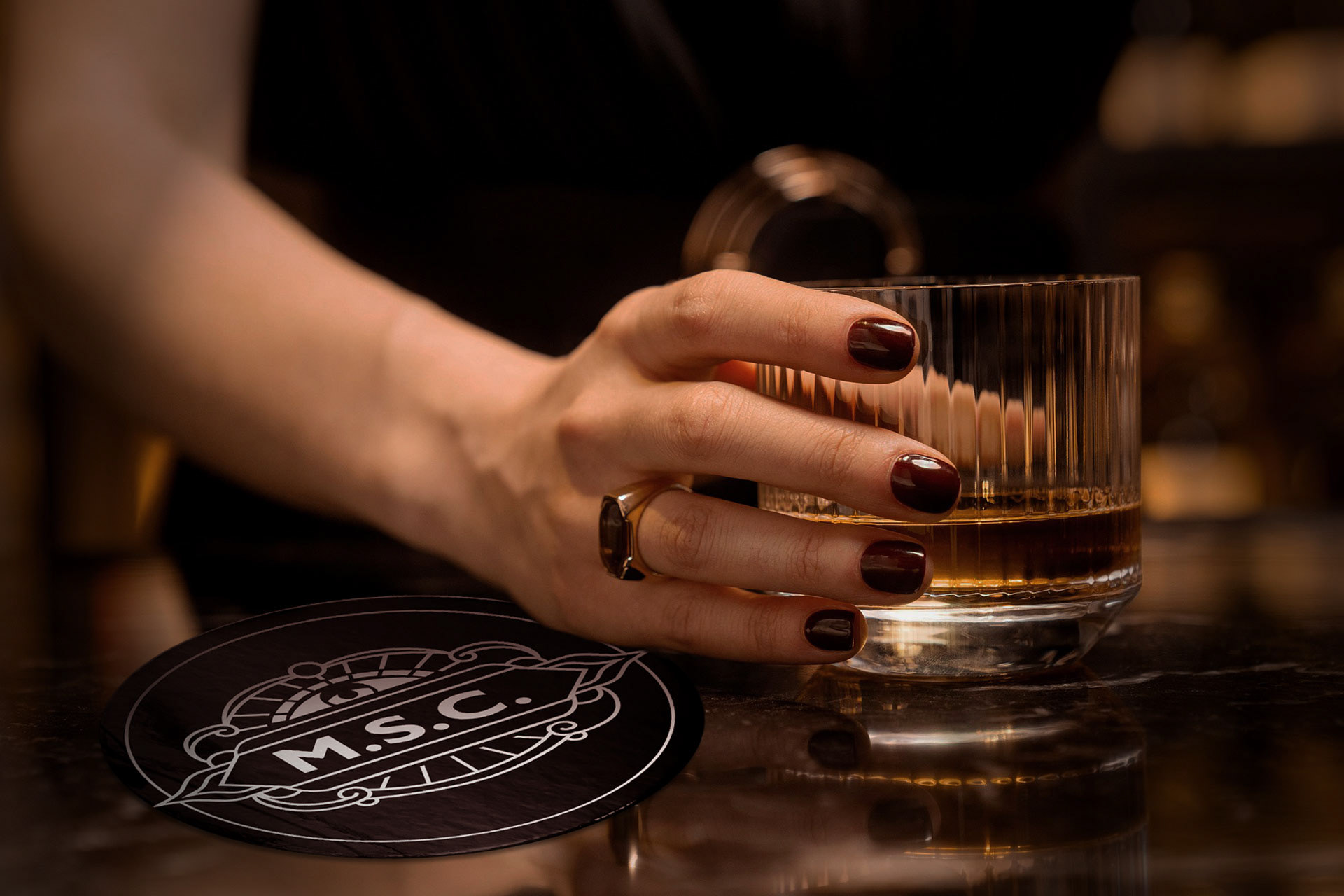 A hand with dark red nails holding a glass of whiskey in a darkly lit bar, next to a black coaster with the Mon Supply Co. logo on it.