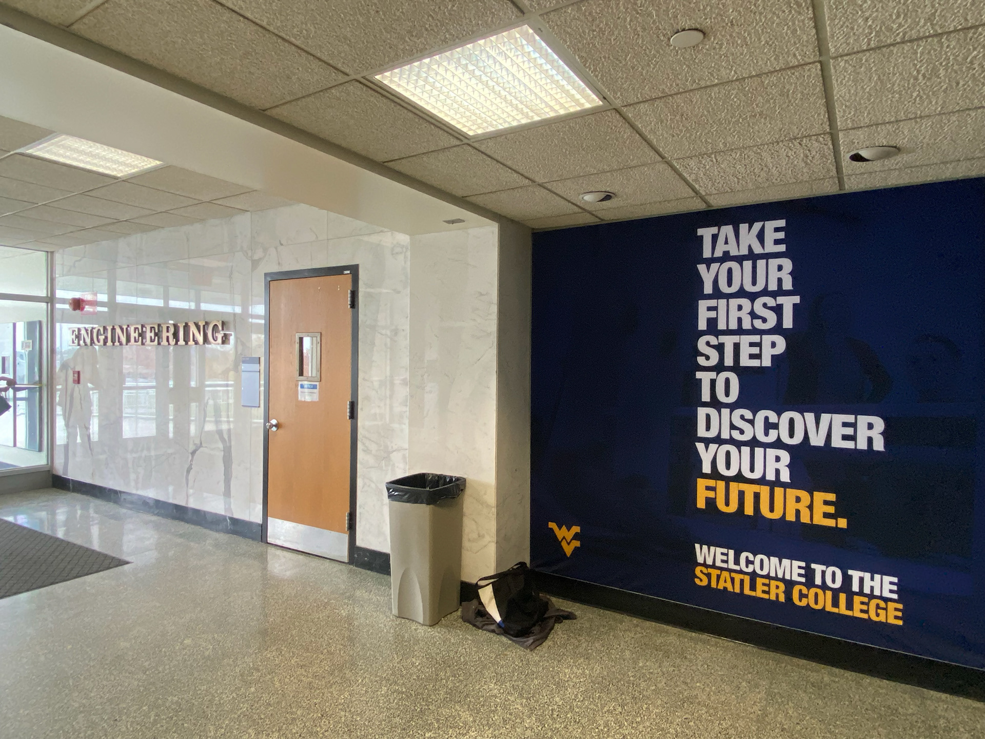The left wall from the entrance to Engineering Sciences building, with a white marble wall with brass 3D letters spelling "ENGINEERING" and a dark navy fabric banner reading "Take your first step to discover your future."