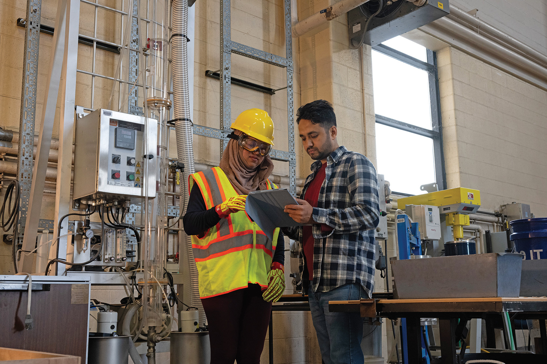 Two students in an industrial setting, one female student wearing full safety vest and hard hat and one male student dressed casually, both examining notes on an iPad