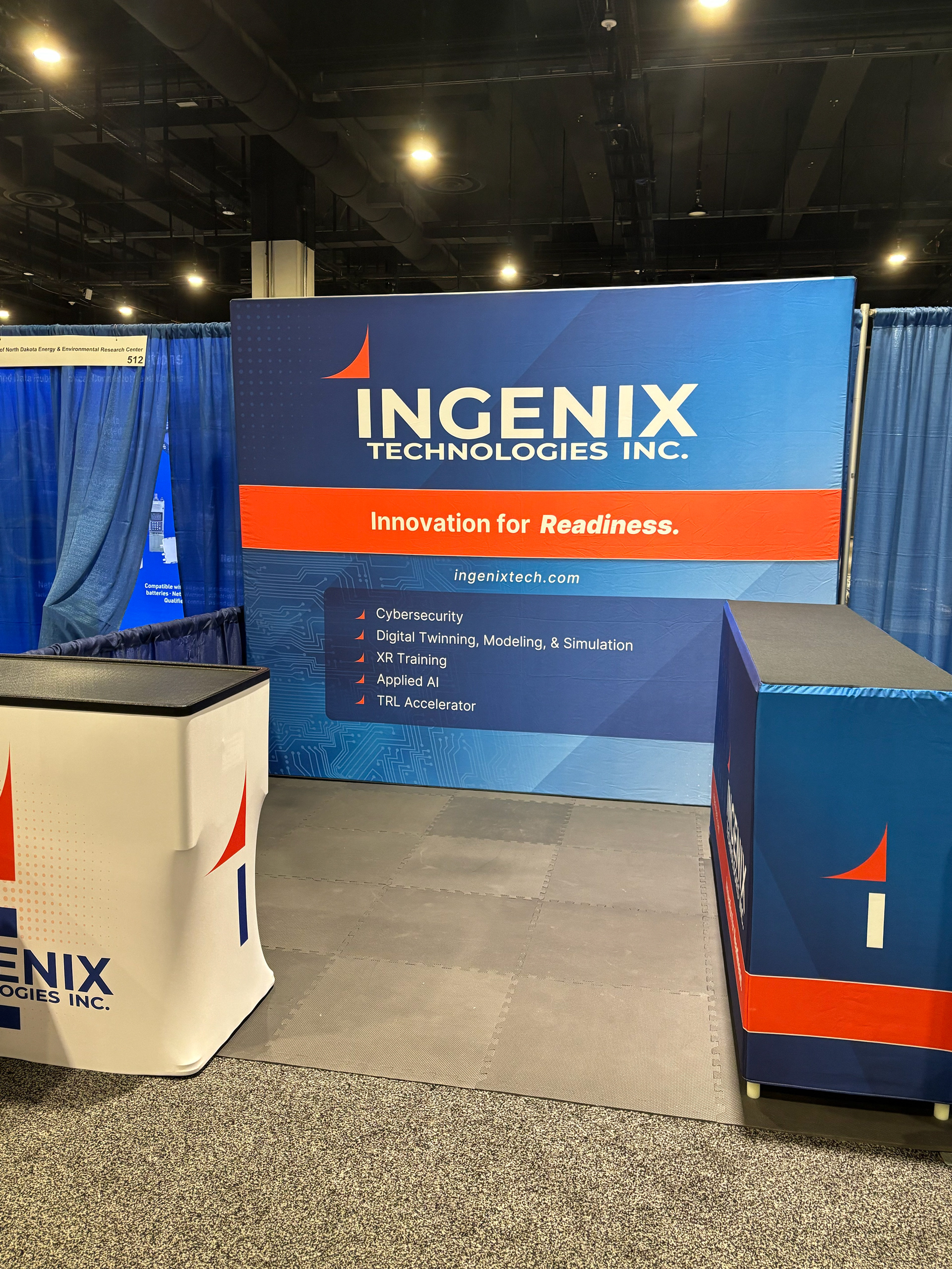 Blue and orange booth backdrop and tables for Ingenix Technologies arranged at a conference. Backdrop reads "Ingenix Technologies Inc.: Innovation for Readiness. ingenixtech.com. Highlighting cybersecurity, digital twinning, modeling, & simulation, XR training, applied AI, and TRL Accelerator."