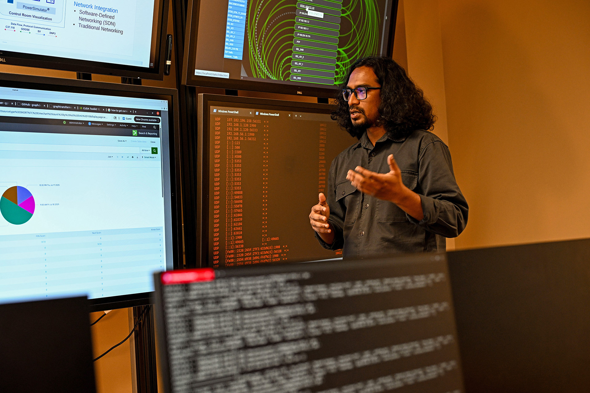 A male student in front of several computer screens explaining complex diagrams and information.