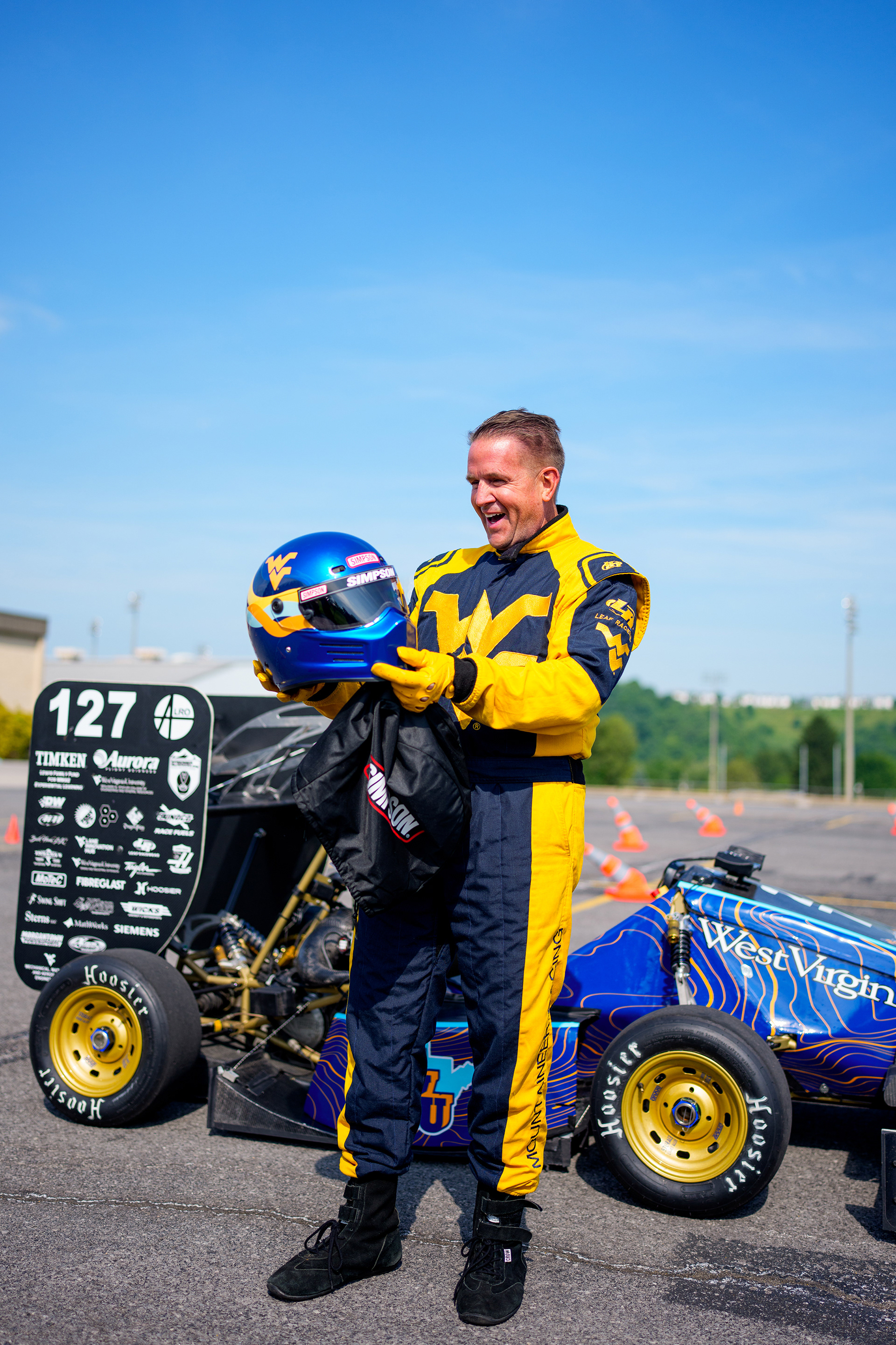 WVU President, Michael Benson, in a blue and gold racing suit holding a blue racing helmet in front of an F1 car on a sunny day.