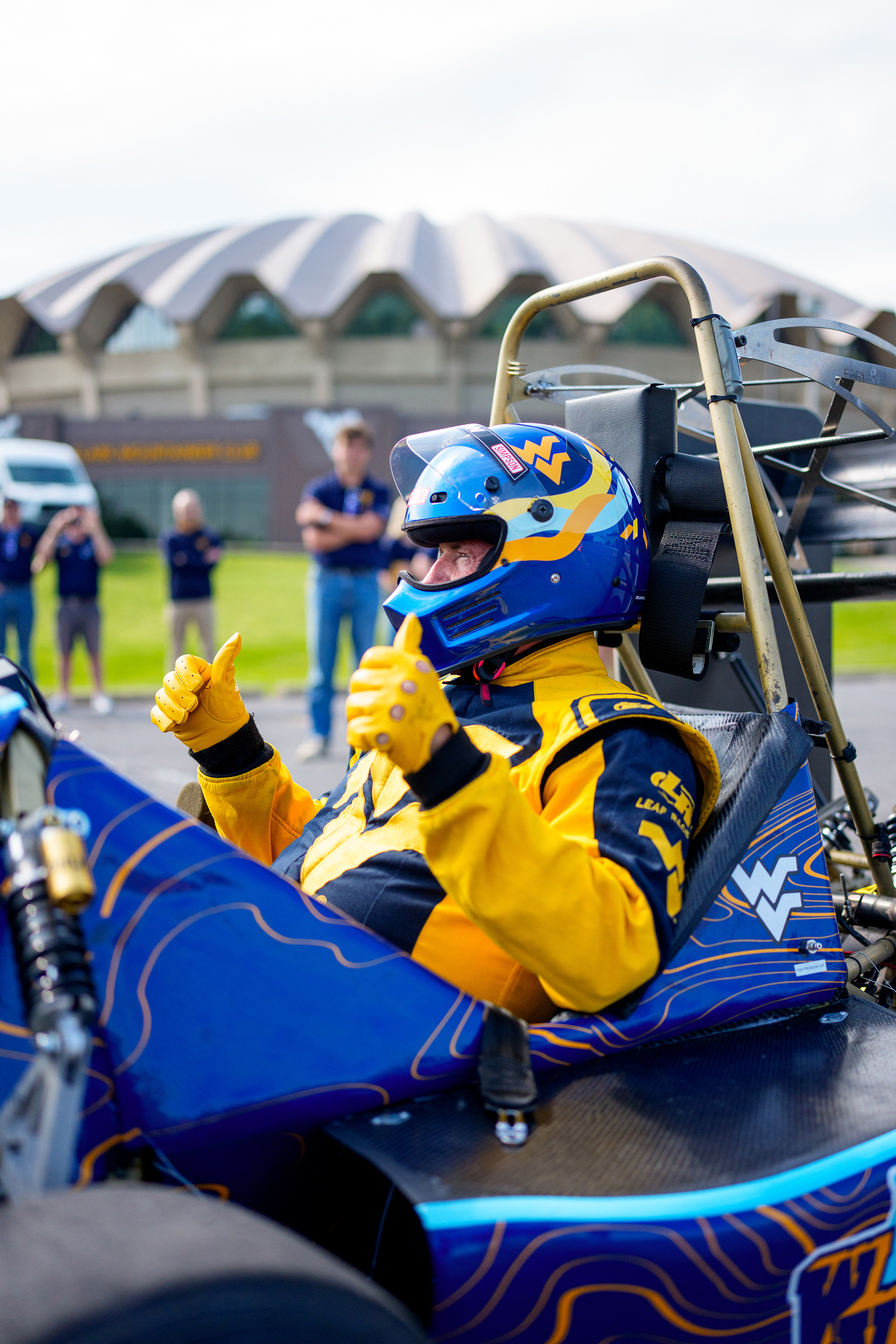WVU President Benson giving a thumbs-up in full facing attire and helmet, while sitting in an F1 racing car in front of the WVU Coliseum.
