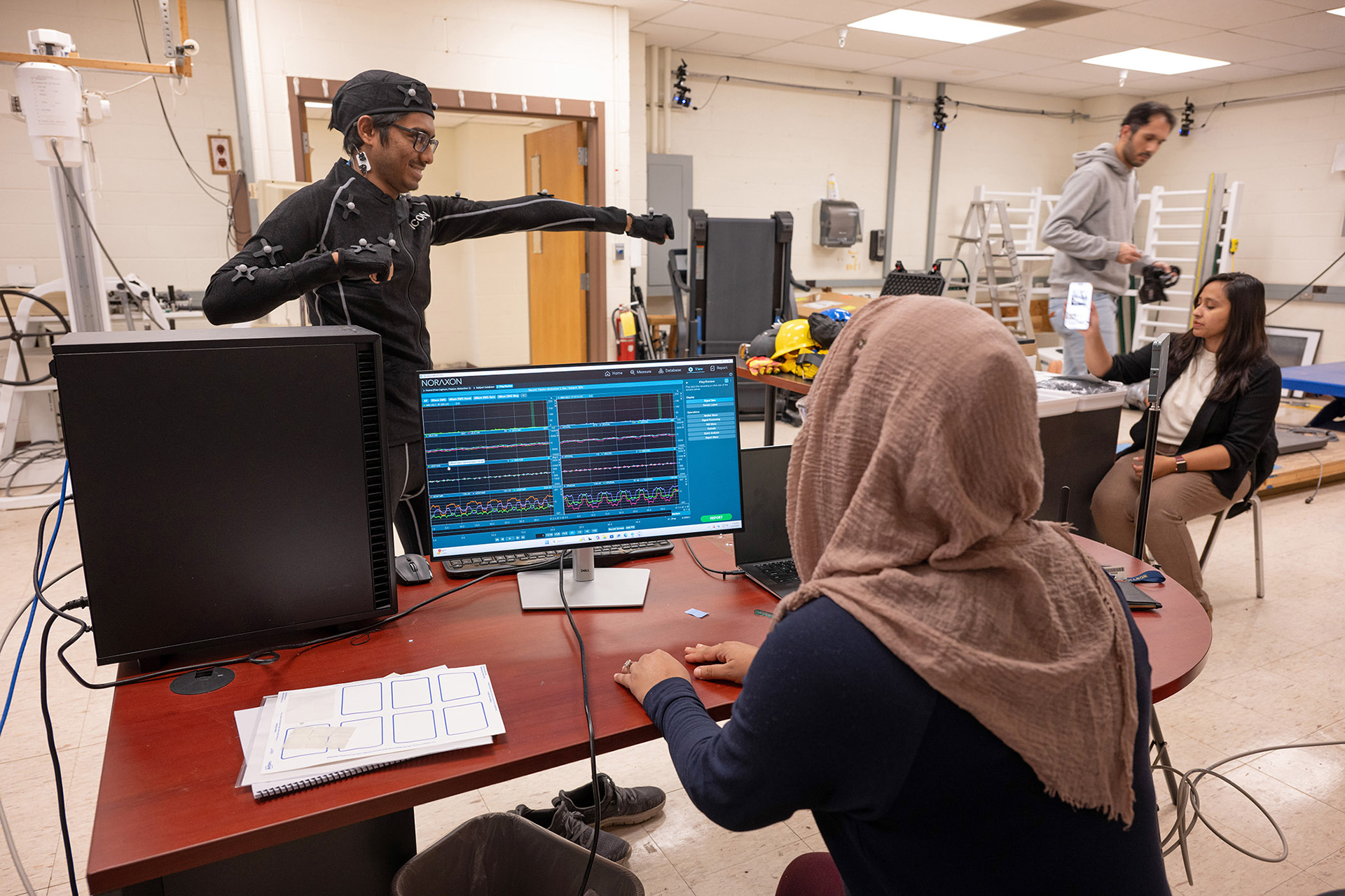 Students examining a male student in full body motion tracking gear, moving his arm forward and making reading appear on the computer monitor.