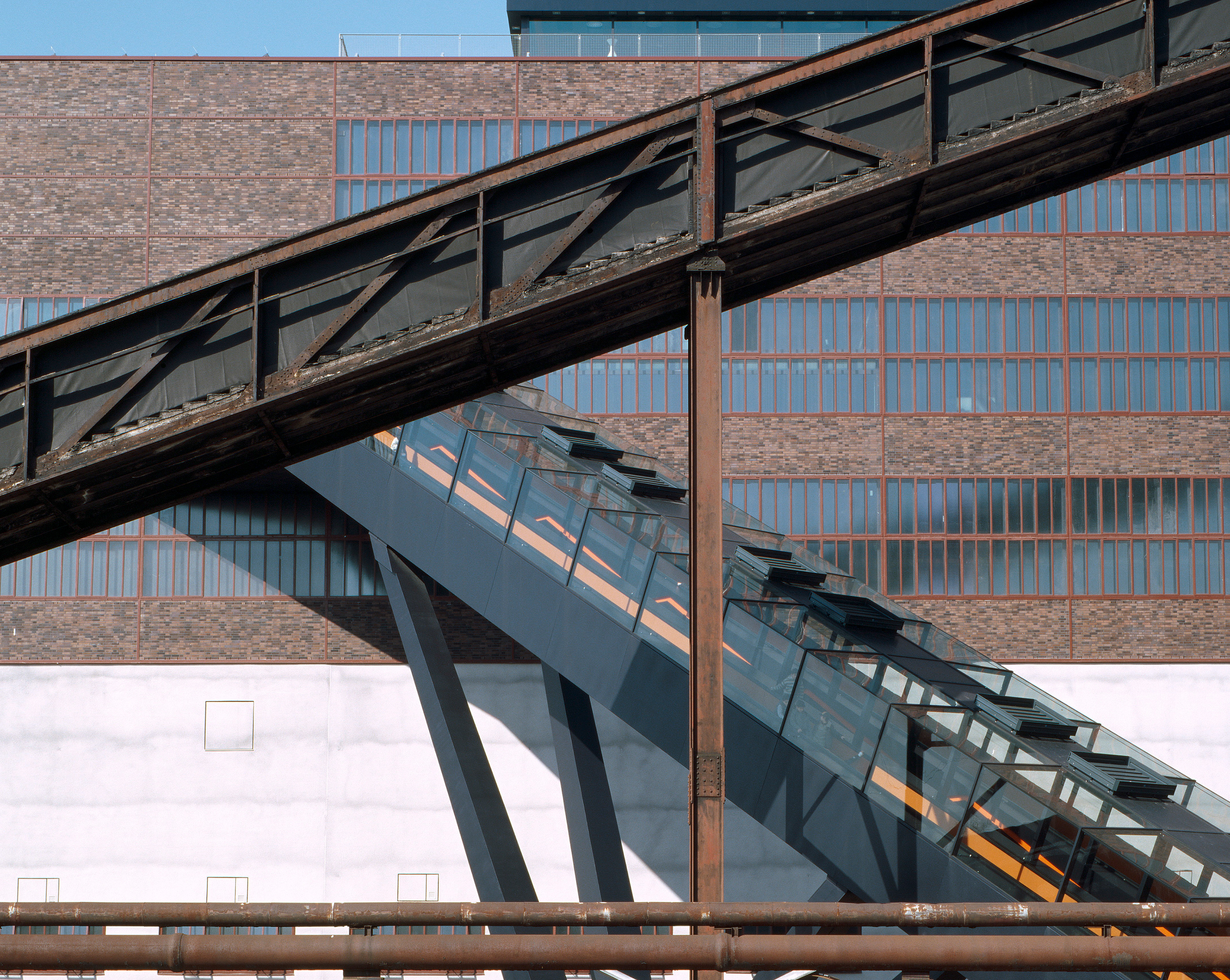 Seitlich-frontale Ansicht der Zeche Zollverein, UNESCO-Weltkulturerbe, mit einem charakteristischen architektonischen Ausschnitt. Sichtbar sind die typischen industriellen Bauelemente: rote Stahlträger, geometrische Strukturen, dunkle Fassadenflächen und die klare, symmetrische Linienführung des ehemaligen Industriekomplexes. Side-frontal view of the Zeche Zollverein UNESCO World Heritage Site, showing a characteristic architectural section. Visible are the iconic industrial elements: red steel beams, geometric structures, dark façade surfaces, and the clean, symmetrical lines typical of the historic coal mining complex.