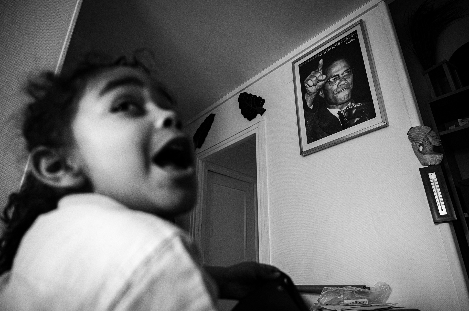 Northern suburbs of Paris, Krissima’s home. March 12, 2026. Krissima's son stands in front of a Malcom X picture which says "Par tout les moyens nécessaires" ("By any means necessary"). 