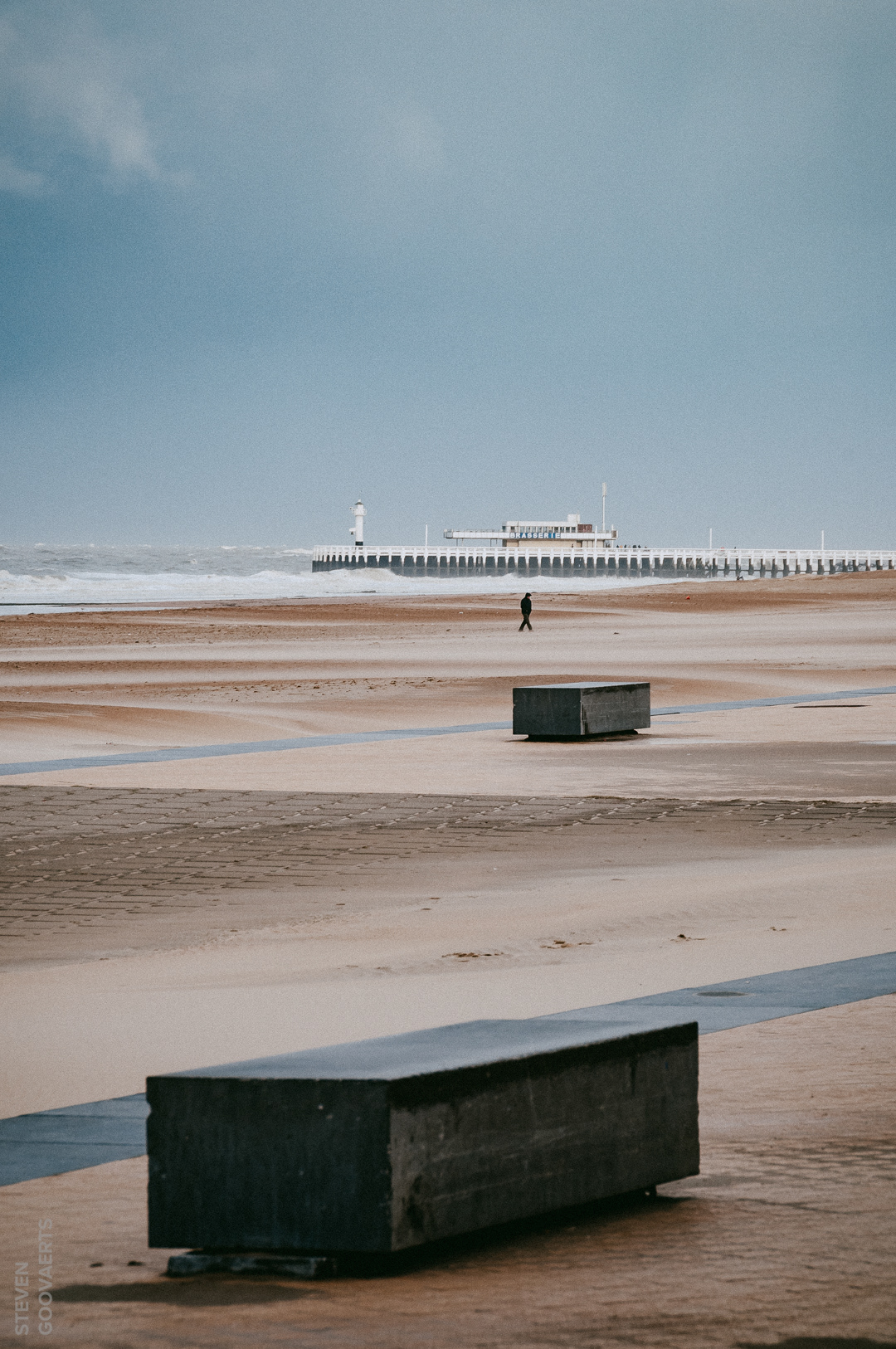Storm aan zee. Eén moedige wandelaar die de beukende wind trotseert. Net als de kriskras geplaatste betonblokken.