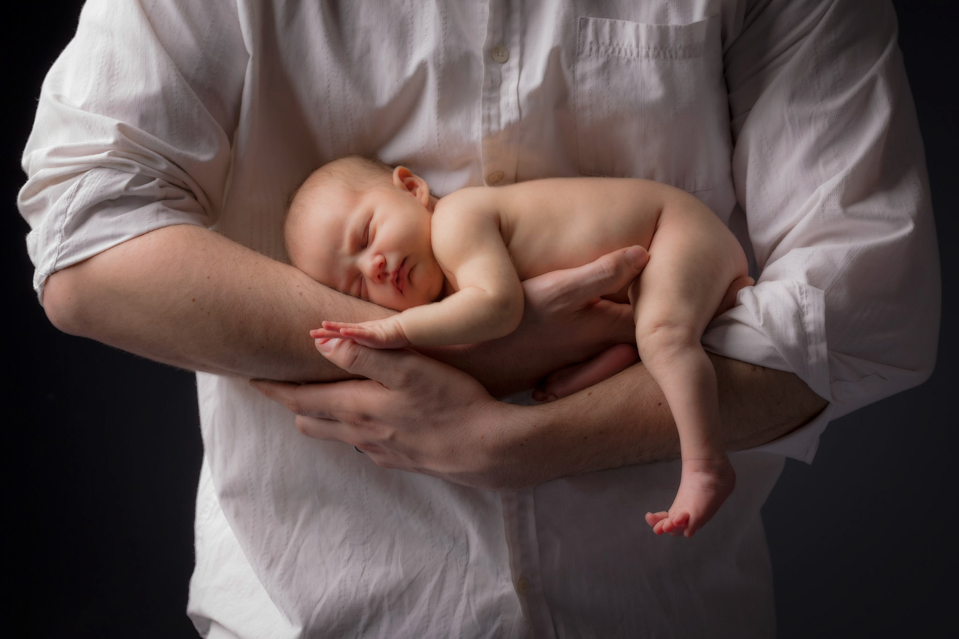 A father cradles his newborn child as she stretches her toes, and touches his thumb for security & comfort.