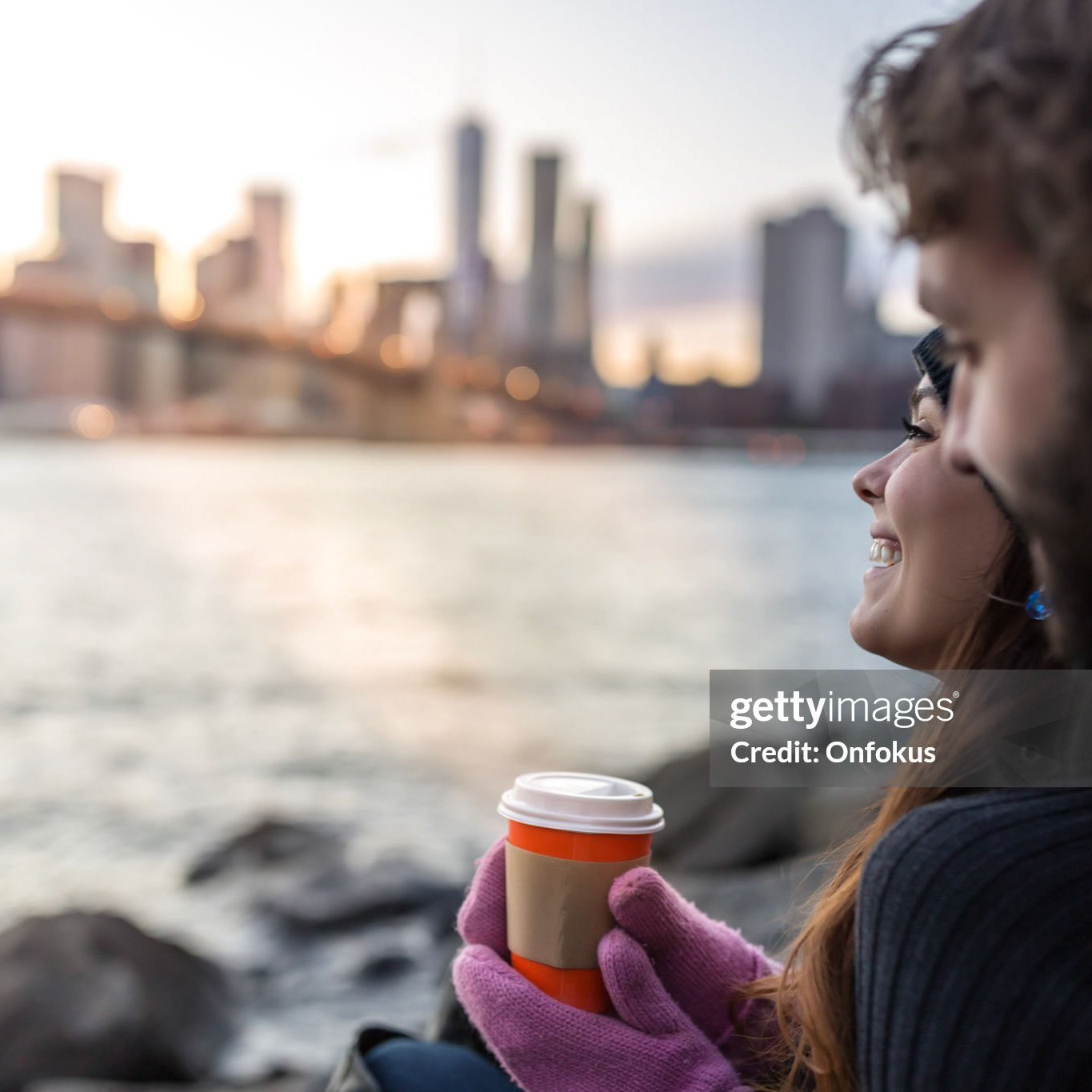 Young couple in love enjoying sunset at Brooklyn Bridge, NYC. The young woman is drinking a coffee while they are having a discussion and smiling.