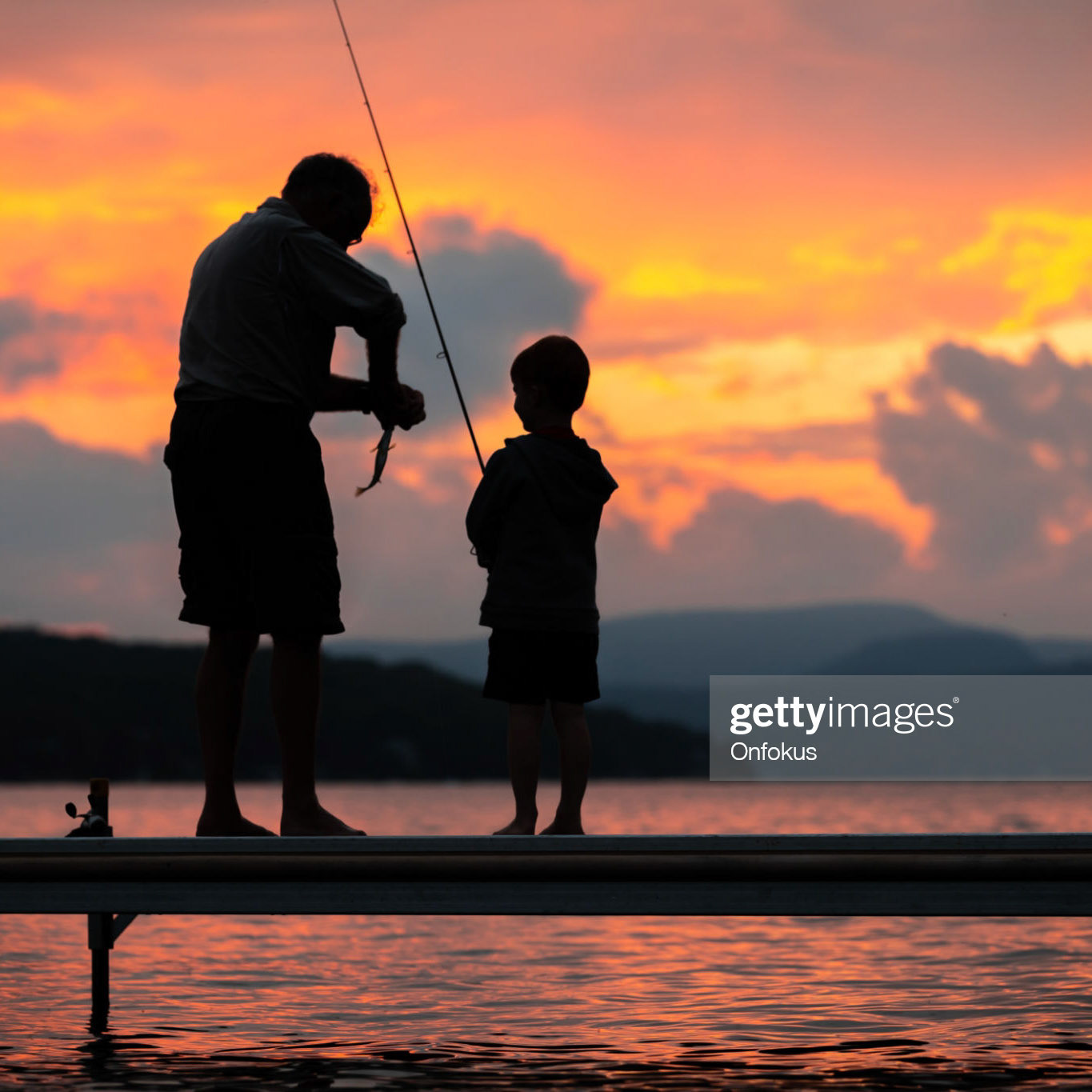 A grandfather is teaching his grandson to fish during summer. They are both standing on the dock. It is a beautiful summer day at sunset. Across the lake, there is a mountain, Quebec, Canada.