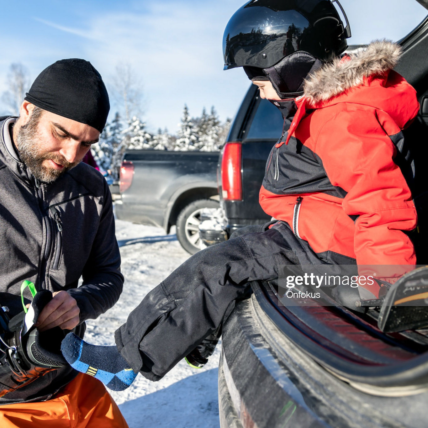 A father and his son are preparing to go ski. They are getting dressed at their car. During the Covid-19 Pandemic, there is a limited access to ski resorts so people have to get dressed and prepare at their car in the parking lot. It is the new normal.