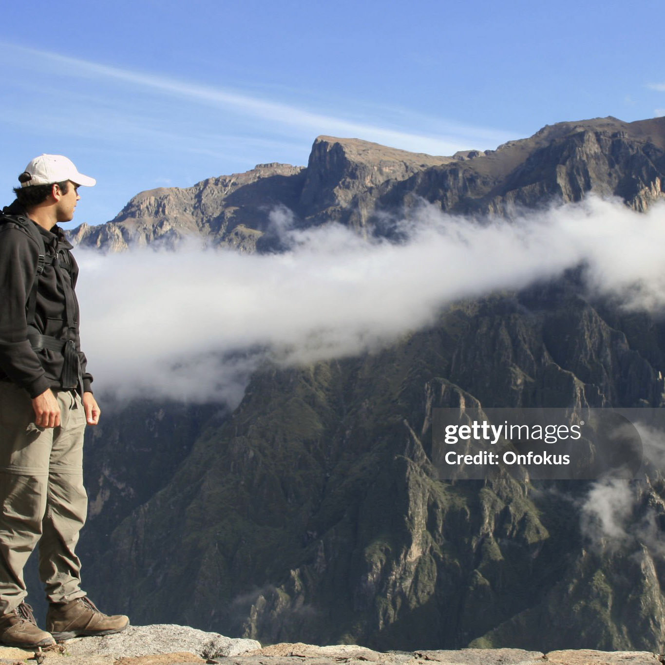 DSLR picture of man hiking with backpack looking at the view inside the Canyon Del Colca, Peru. The sky is blue and the backpacker is above the clouds.
