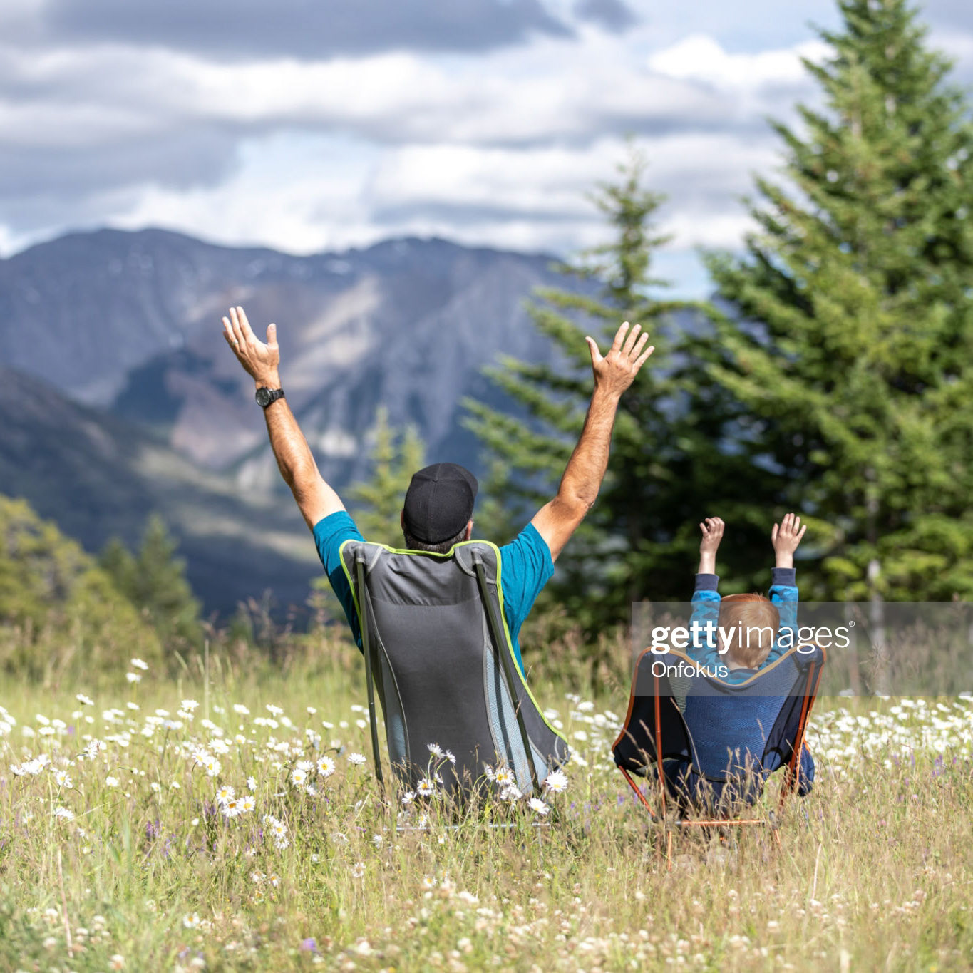 Father and Son sitting on camping chairs with arm raised and looking at mountains in Banff National Park in Summer.