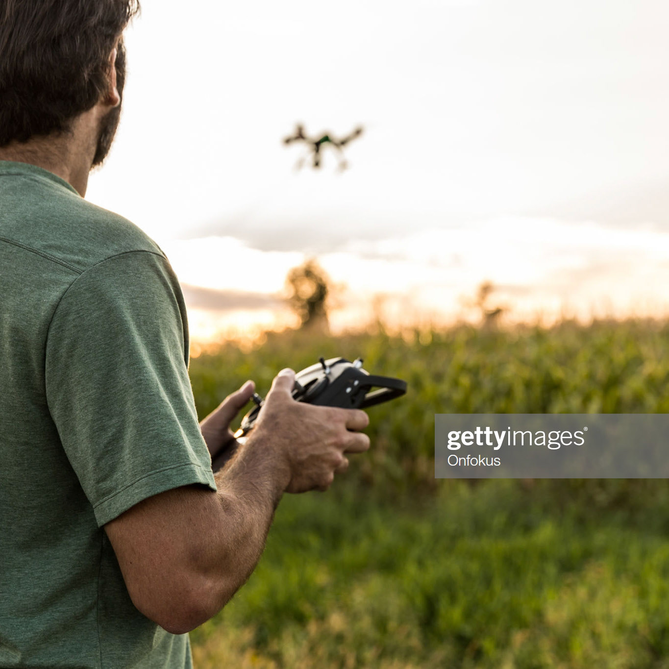 DSLR picture of a man flying a drone over a field by a beautiful summer sunset. The man is looking at the drone.