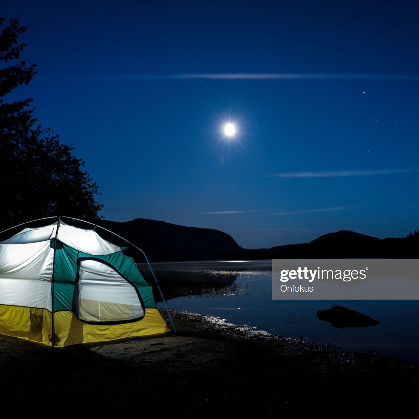 Campsite, tent close to lake at night. You can see the reflection of the moon on the lake. There is a light in the tent. Mont-Tremblant, Quebec, Canada