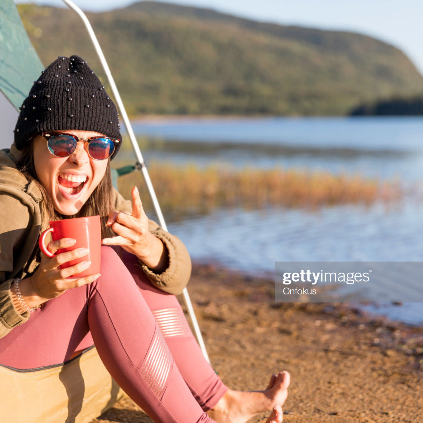 A young women is drinking a coffee by the lake in camping. She is sitting in her tent and contemplating the view. She is wearing warm clothing.