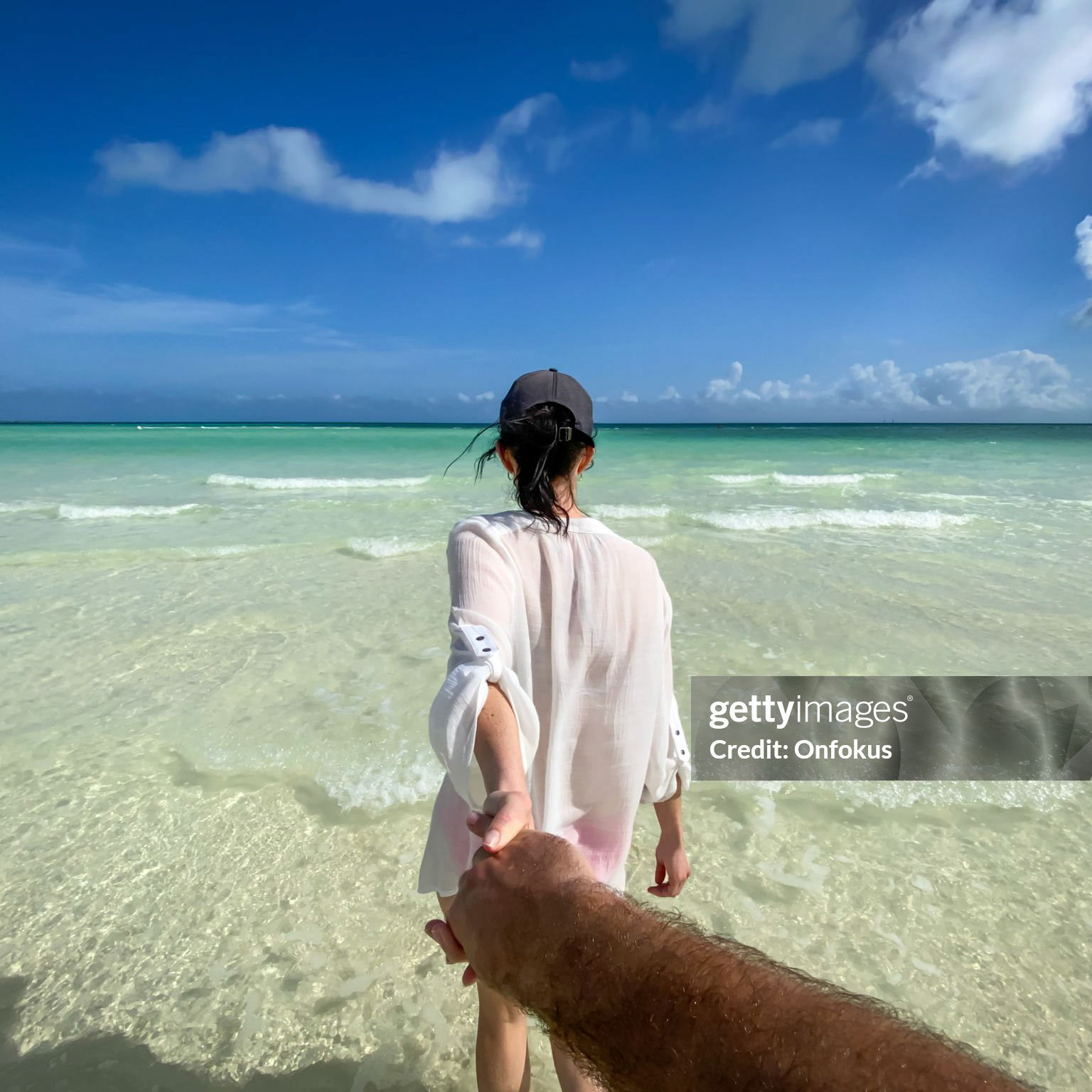 Follow me concept - A beautiful woman is holding her husband’s hand in the ocean. The photography is taken from the man’s point of view, personal perspective. They are walking on a beautiful tropical beach in Cuba, towards the turquoise colored Caribbean sea. They are in love and it is their honeymoon.