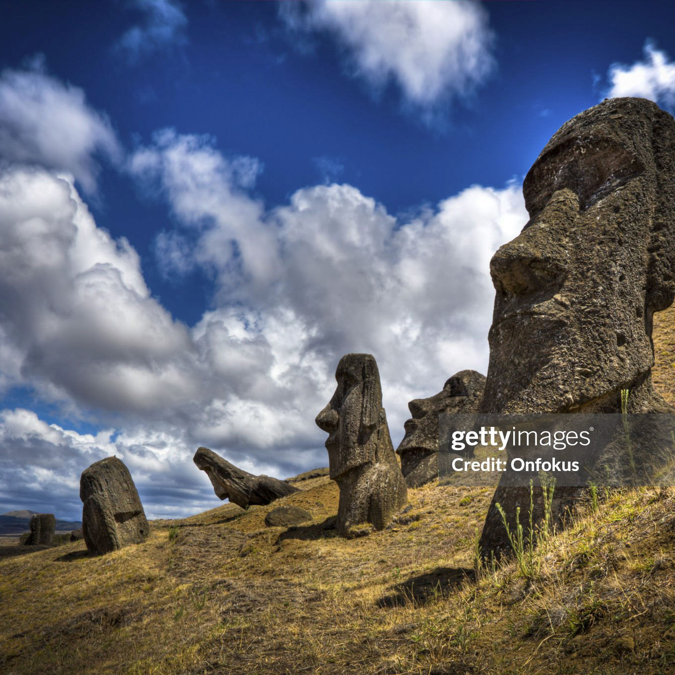 DSLR picture of Moais at the Easter Island in Chile. Moais are human figures carved by the Rapa Nui people in rock. One Moais is in the foreground and four in the background. The sky is mostly sunny with clouds.