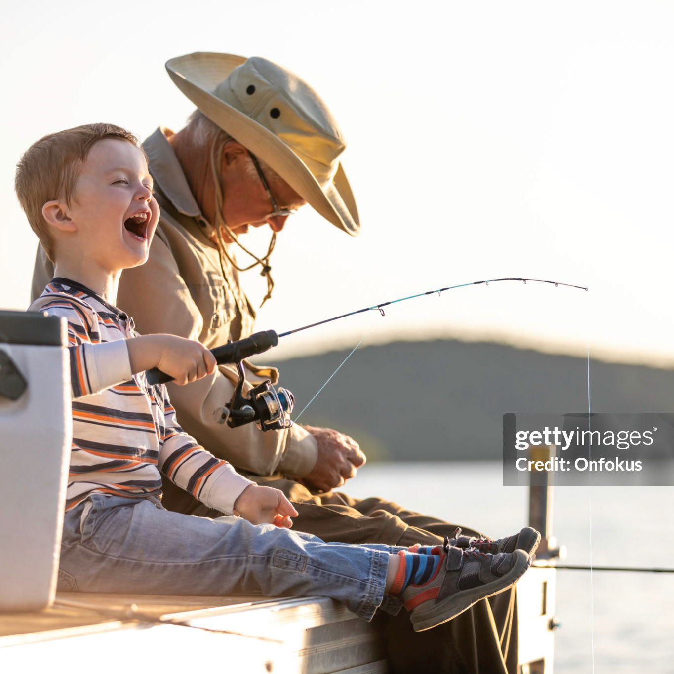 A grandfather is teaching his grandson to fish during sunset in summer. They are both sitting on the dock and laughing. It is a beautiful summer day. Across the lake, there is a mountain.