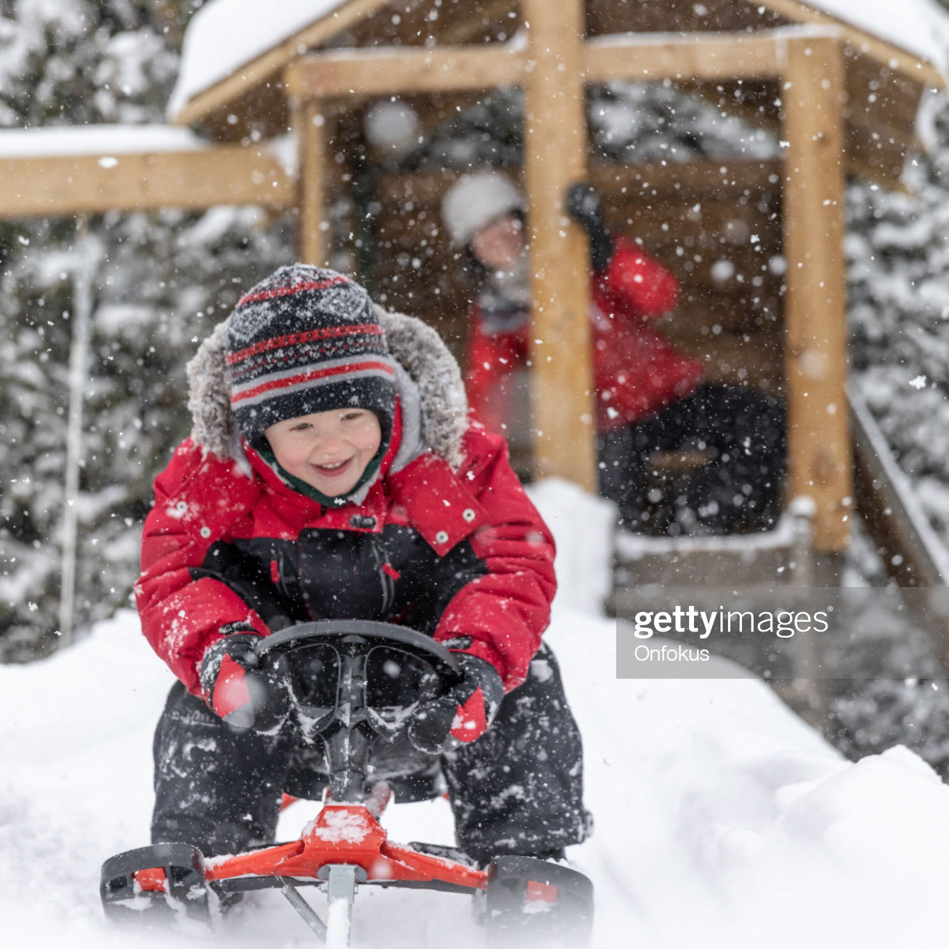 A little redhead boy is sledding on a red and black snowracer (sleigh) on the snow, outdoors in winter. He is home, in his backyard. His mother is pushing him, looking at him and smiling in the background.