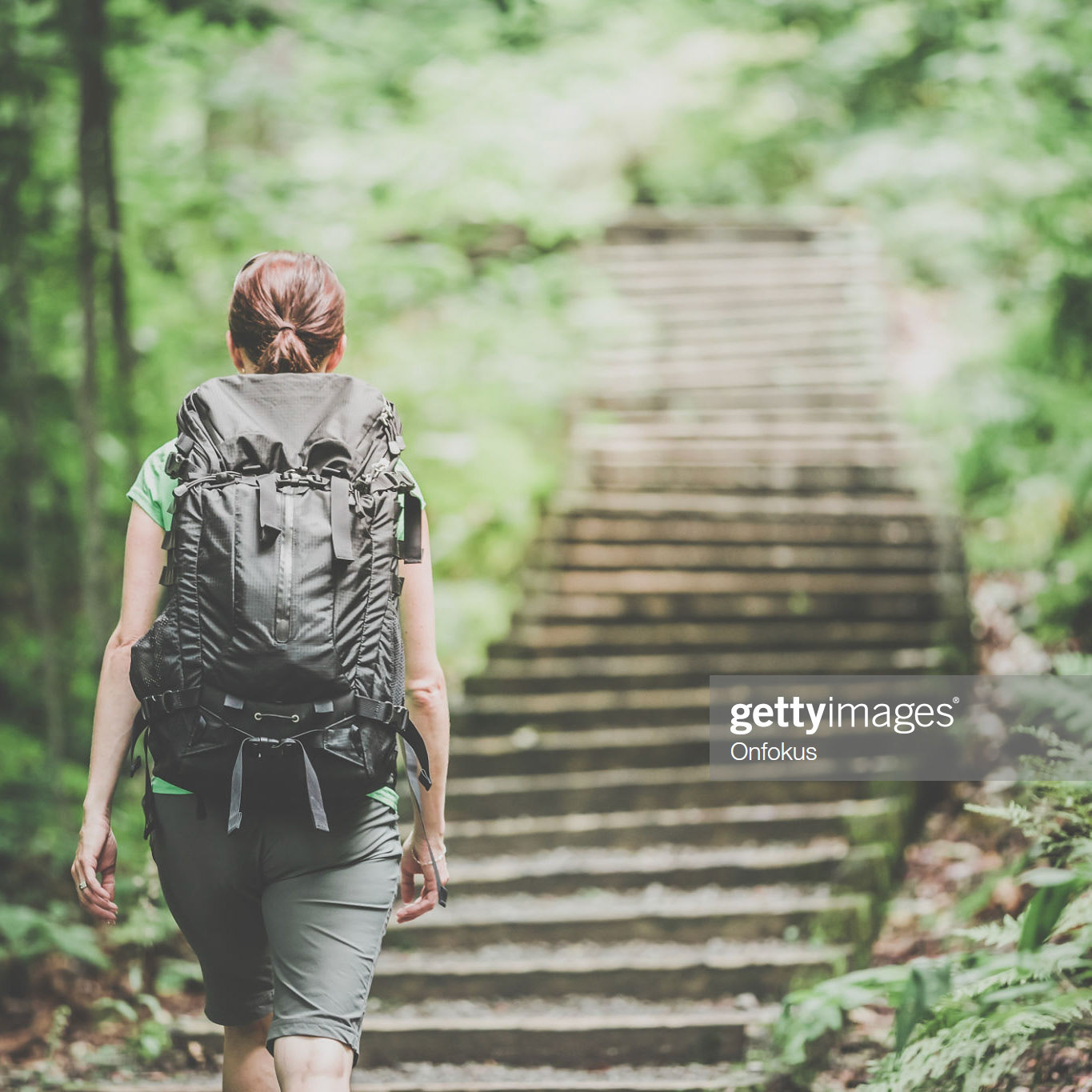 DSLR picture of a woman hiking or trekking with backpack on a footpath in forest on a nice day of summer. There is wood and rock stairs to climb in front of her.