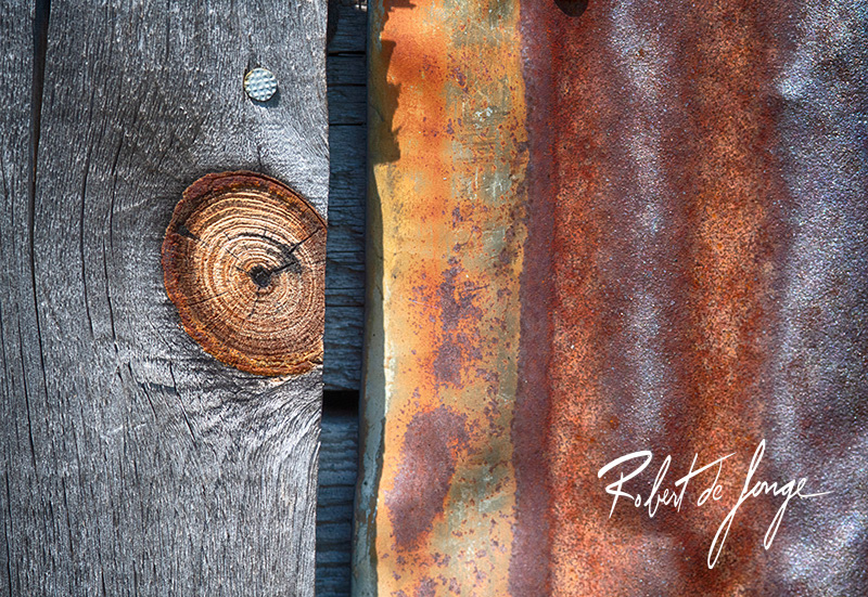 Wood siding on a farm outbuilding displays a knot that borders some rusty corrogated siding