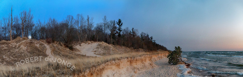 A panorama of the shoreline at Hoffmaster State Park in the evening. The Lake tree at the water's edge with ice ridges around it. The moon is rising over the low dune bluffs