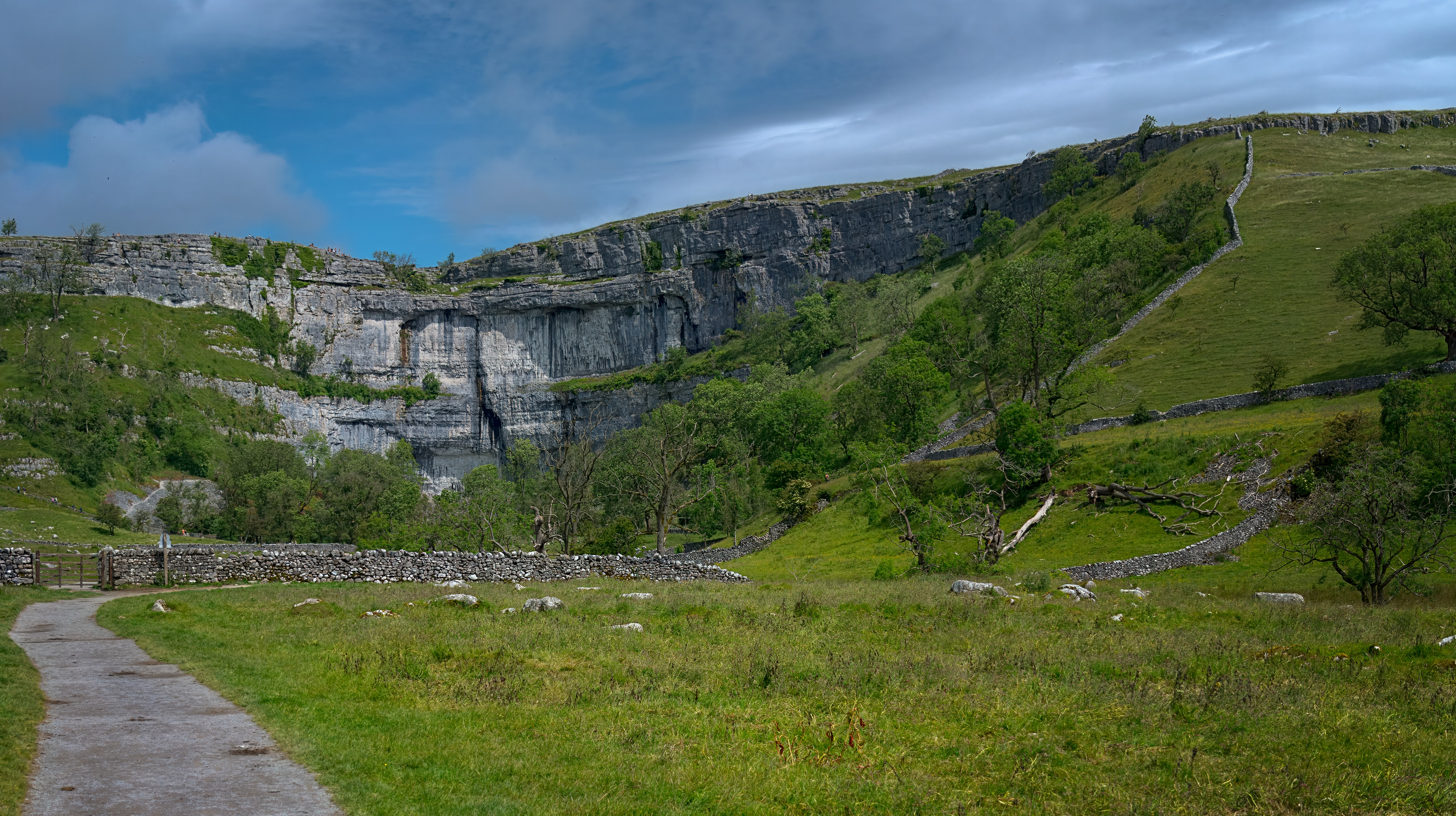 A fine-art landscape photograph showing a vast, curved limestone cliff towering over a lush green valley, featuring a winding dry stone wall and a quiet walking path beneath a dynamic, partly cloudy blue sky.