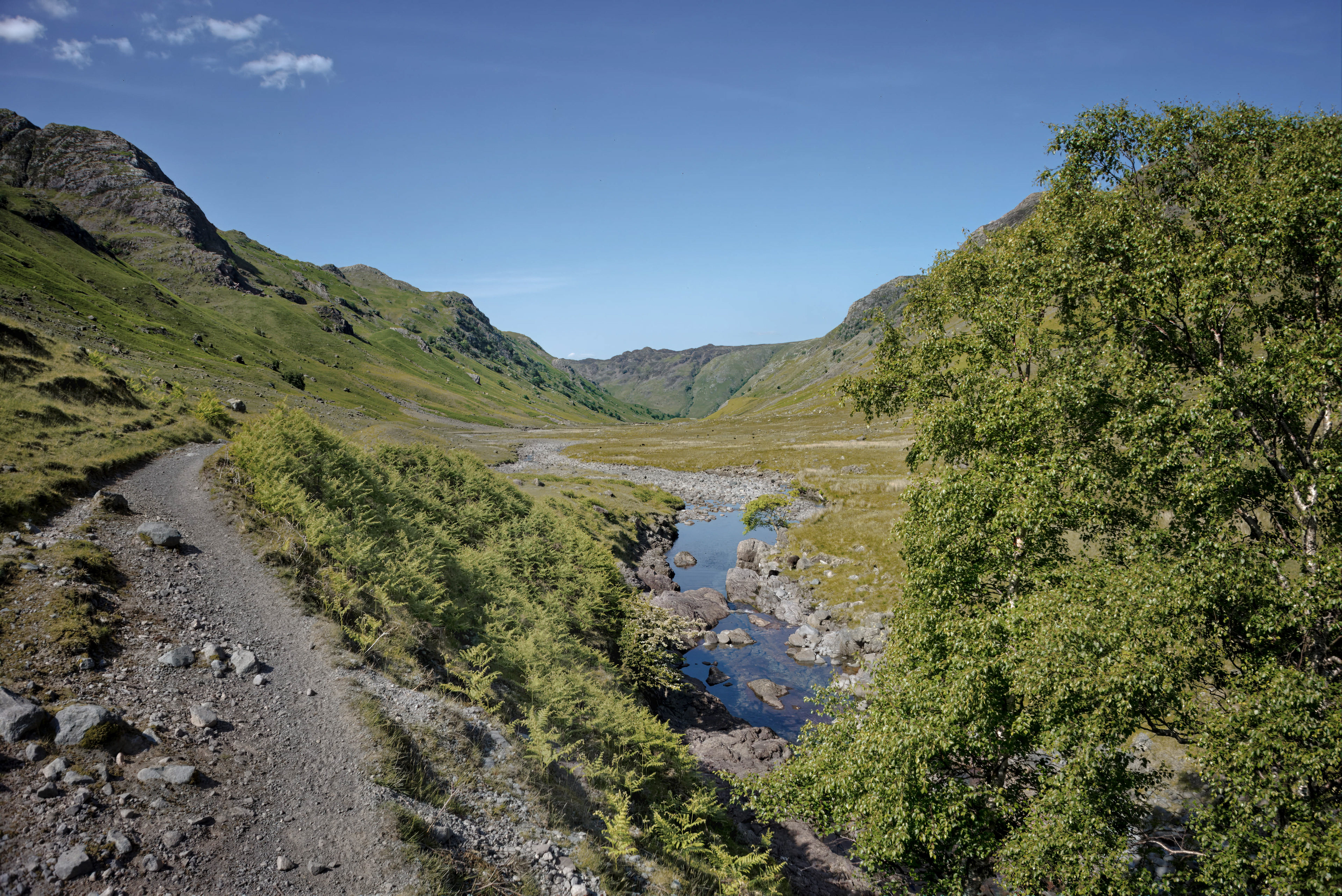 A sweeping landscape view of a vast, green valley featuring a rugged gravel path on the left, a shallow rocky stream winding through the center, and steep, verdant hills rising under a clear blue sky.