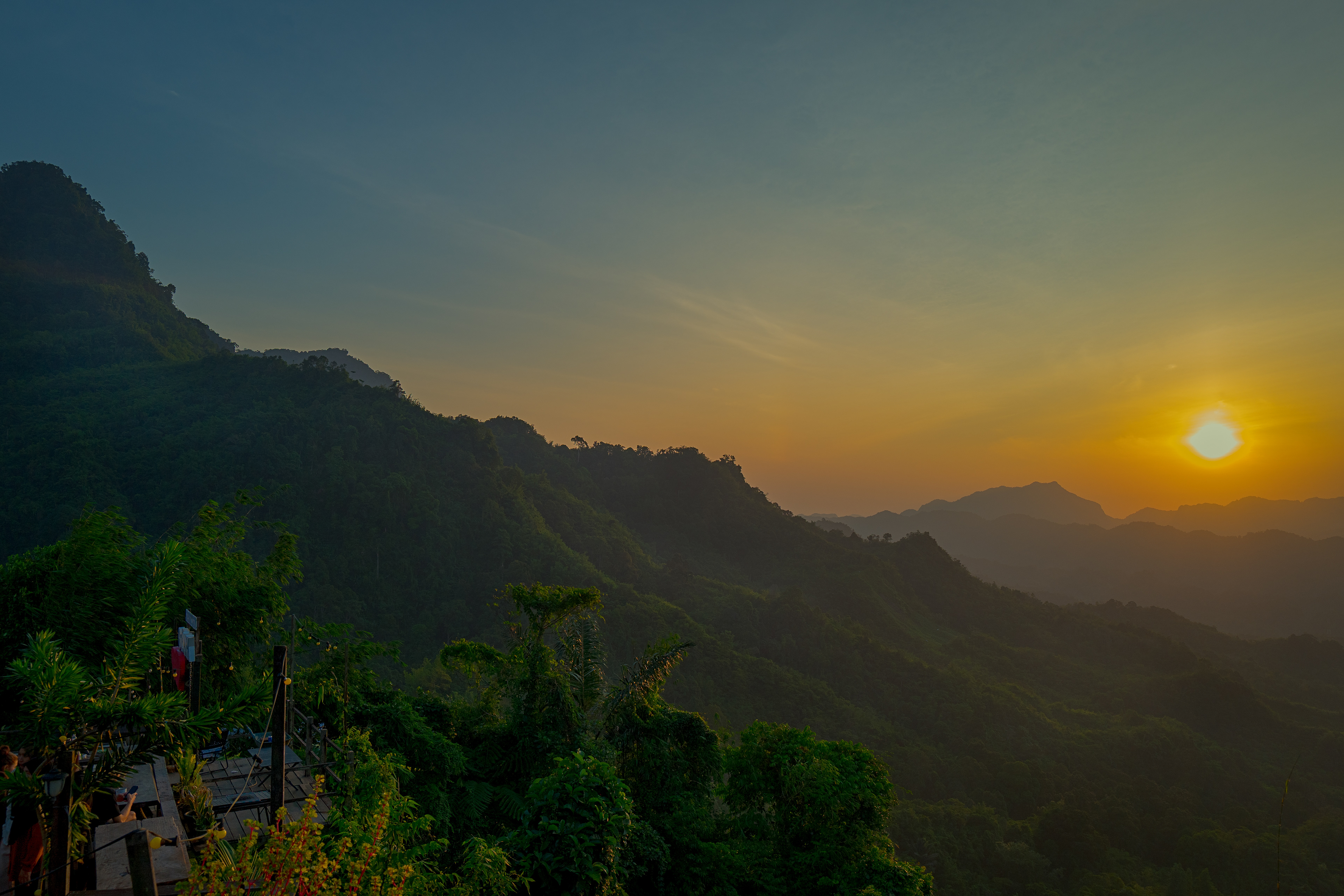 A panoramic sunrise over rainforest-covered mountains, with golden morning light illuminating layers of misty ridgelines beneath a clear sky. Lush tropical vegetation fills the foreground as the first light of day reveals the dramatic landscape at the Kalimantan–Malaysia Border near Kampong Sapit, Padawan.