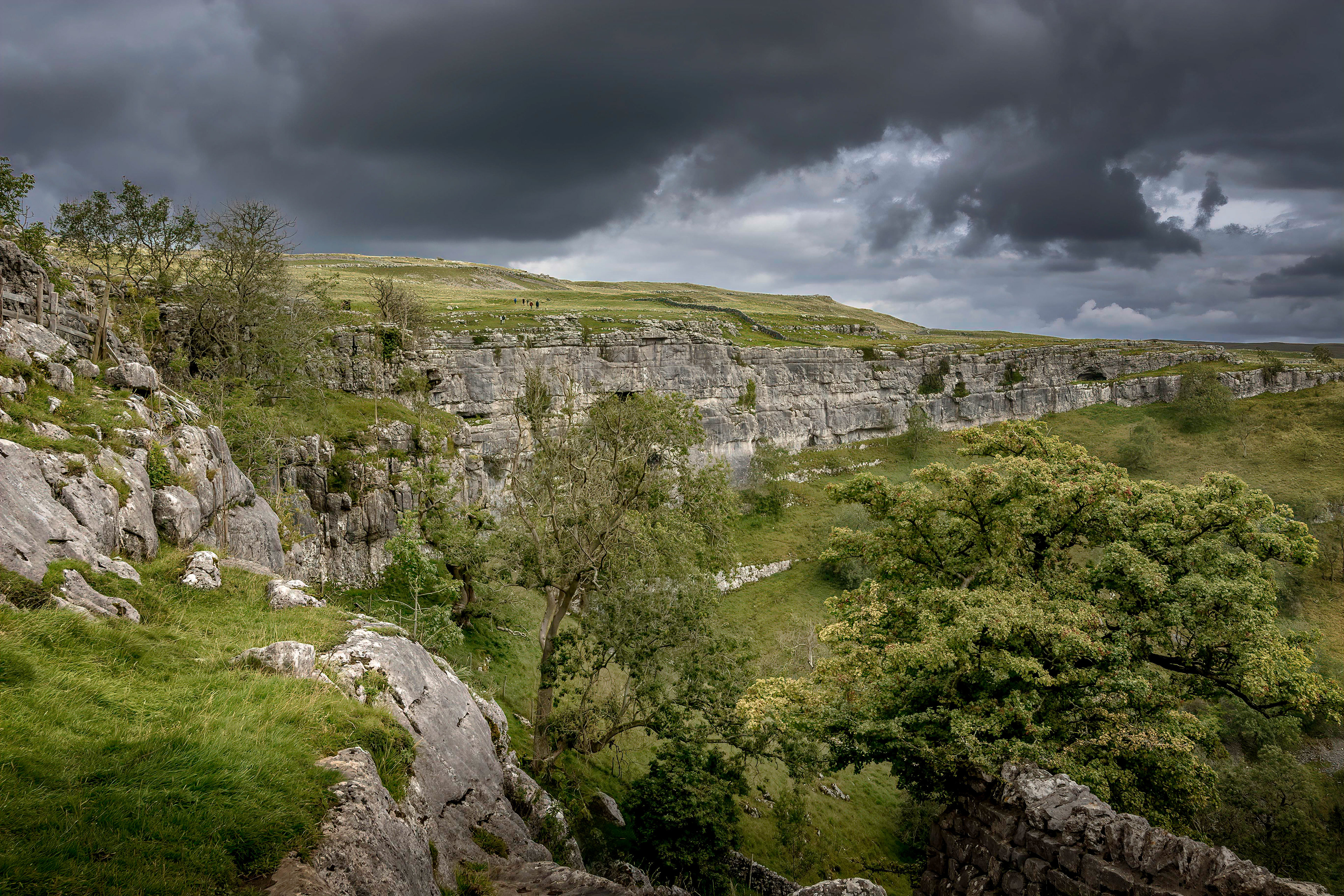 Massive limestone cliffs of Malham Cove rising beneath dark storm clouds, with green valleys, trees, and distant figures across the Yorkshire Dales landscape.