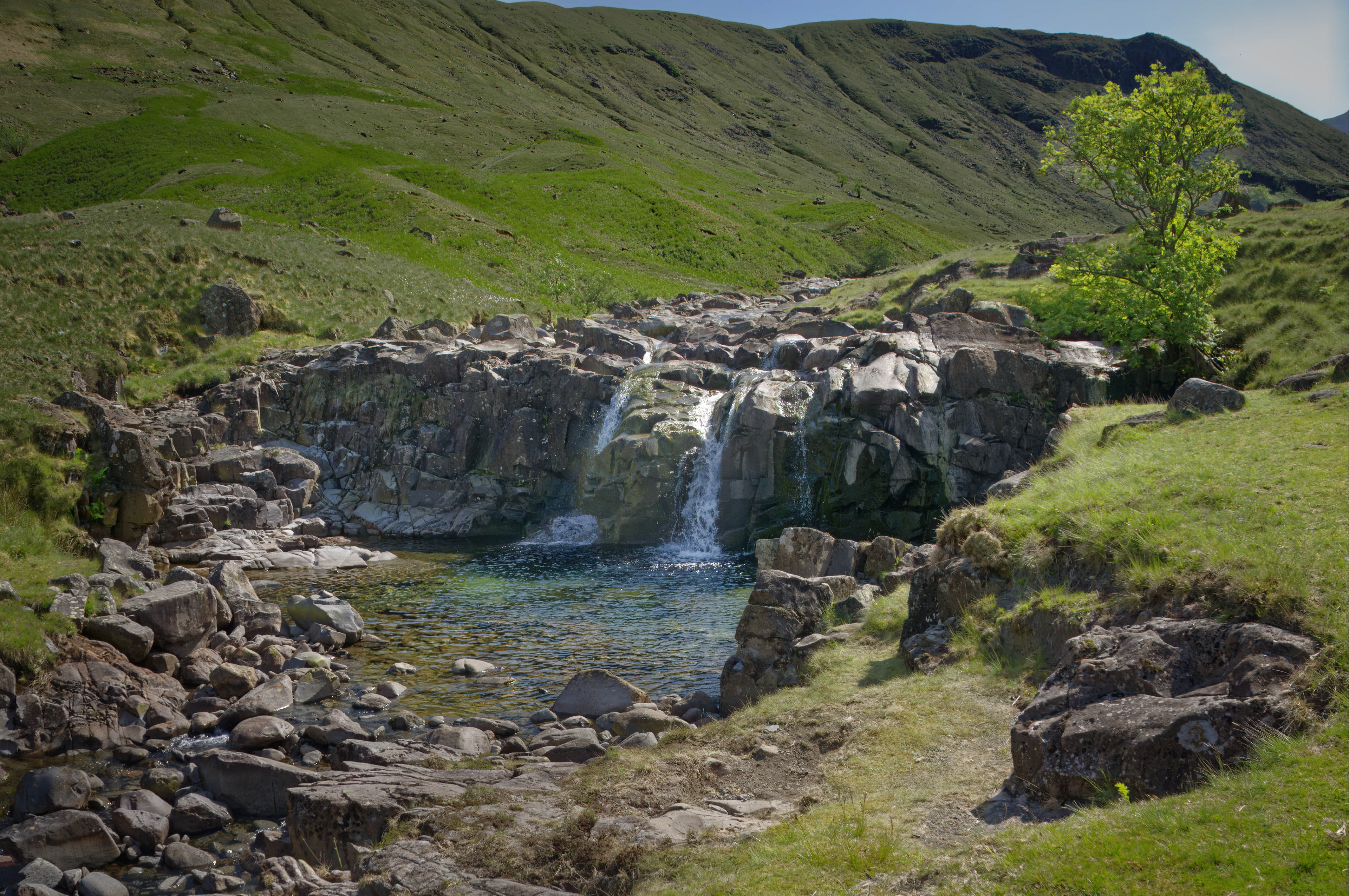 A fine-art landscape photograph of a rocky waterfall cascading into a shallow, clear pool, surrounded by vibrant green, sunlit grassy fells and a single leafy tree under a bright sky.