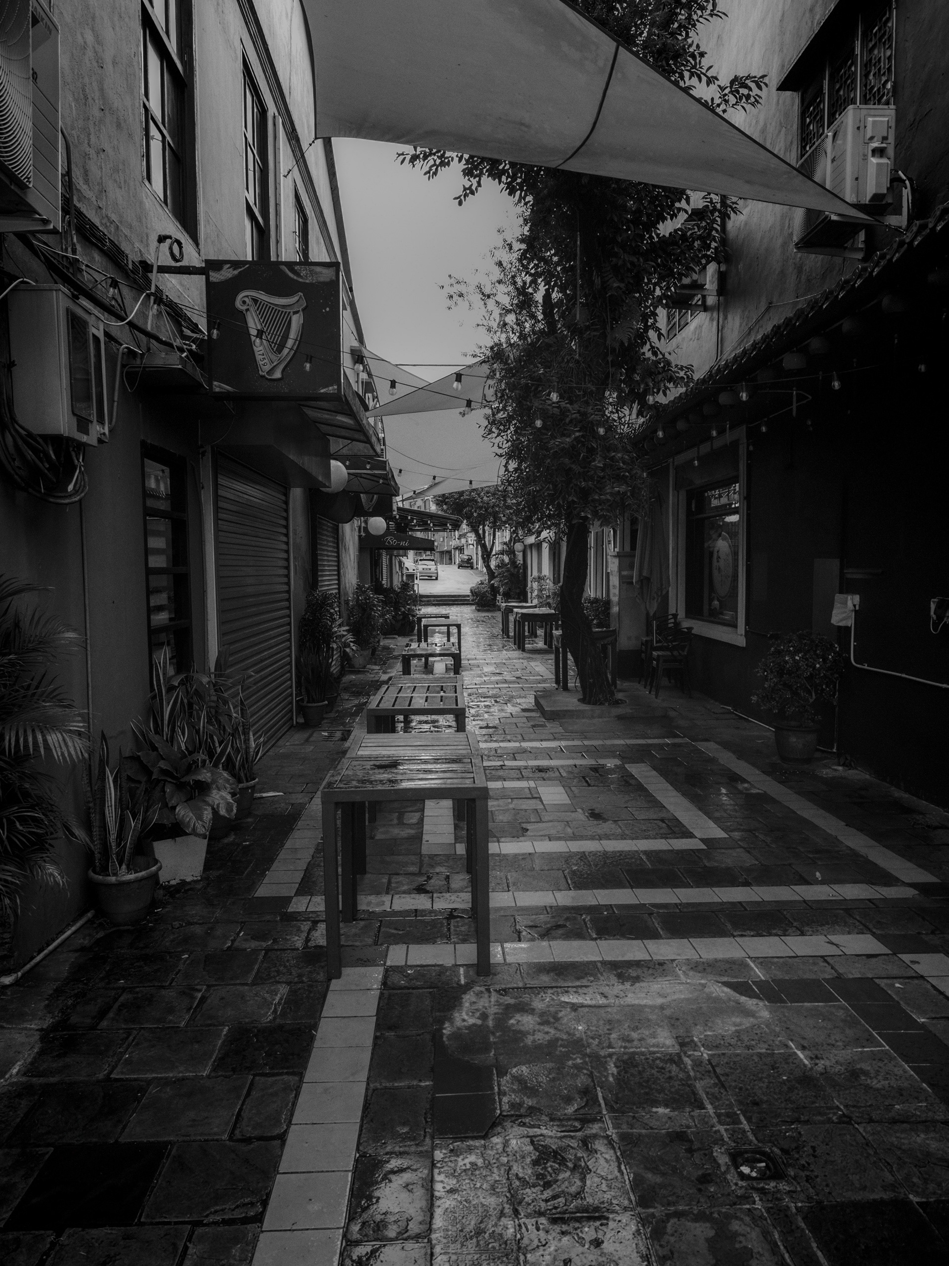 A black-and-white photograph of a narrow urban alley lined with café tables, potted plants, and weathered buildings. Overhead fabric canopies and string lights frame the scene, while rain-soaked paving stones reflect soft light, creating a quiet, atmospheric composition that draws the eye toward the distant street in Kuching, Sarawak.