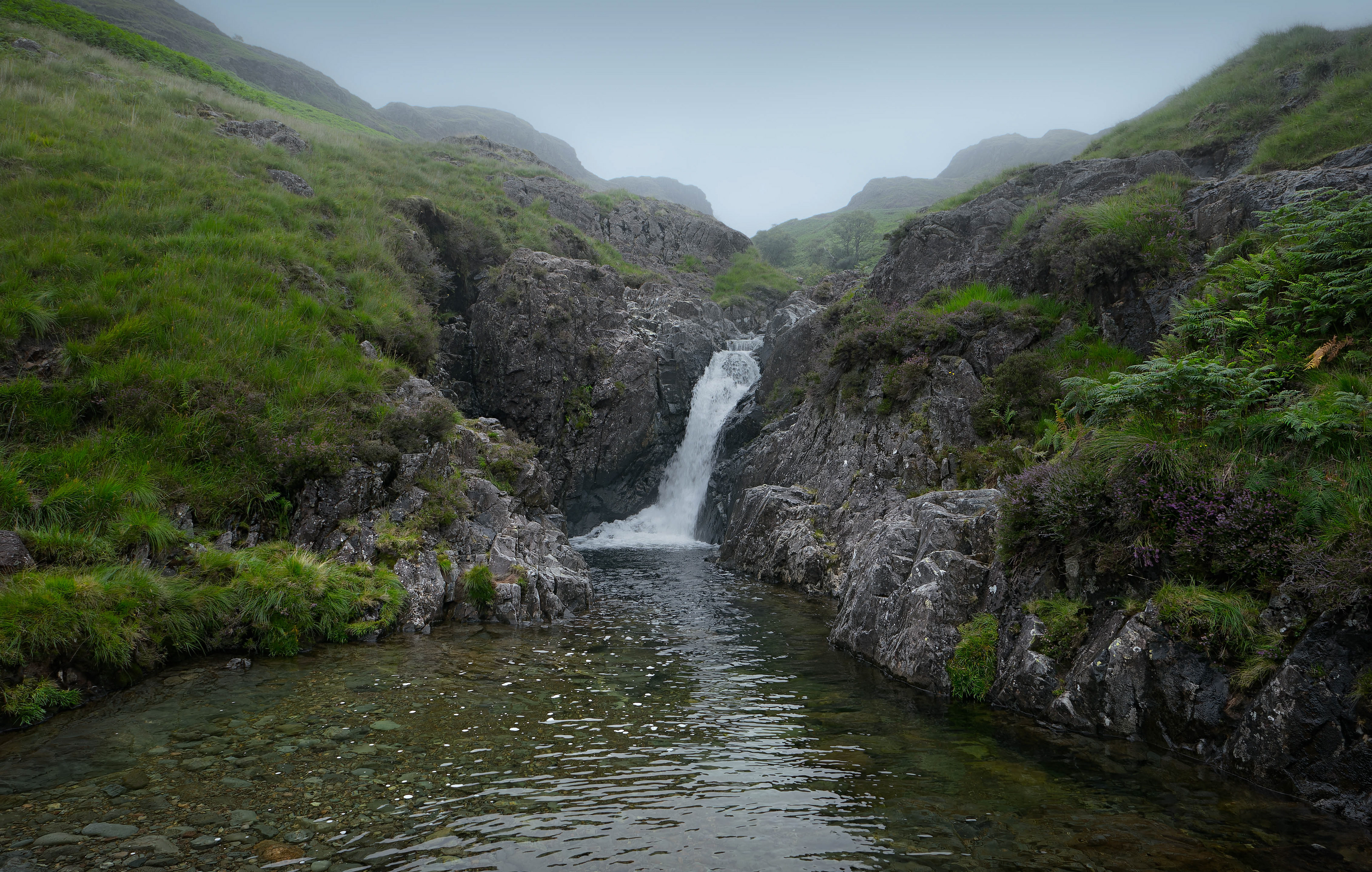 Misty, atmospheric view of Upper Esk Falls in Eskdale, with a waterfall cascading between dark craggy rocks into clear pools, surrounded by lush green hills under a pale overcast sky.