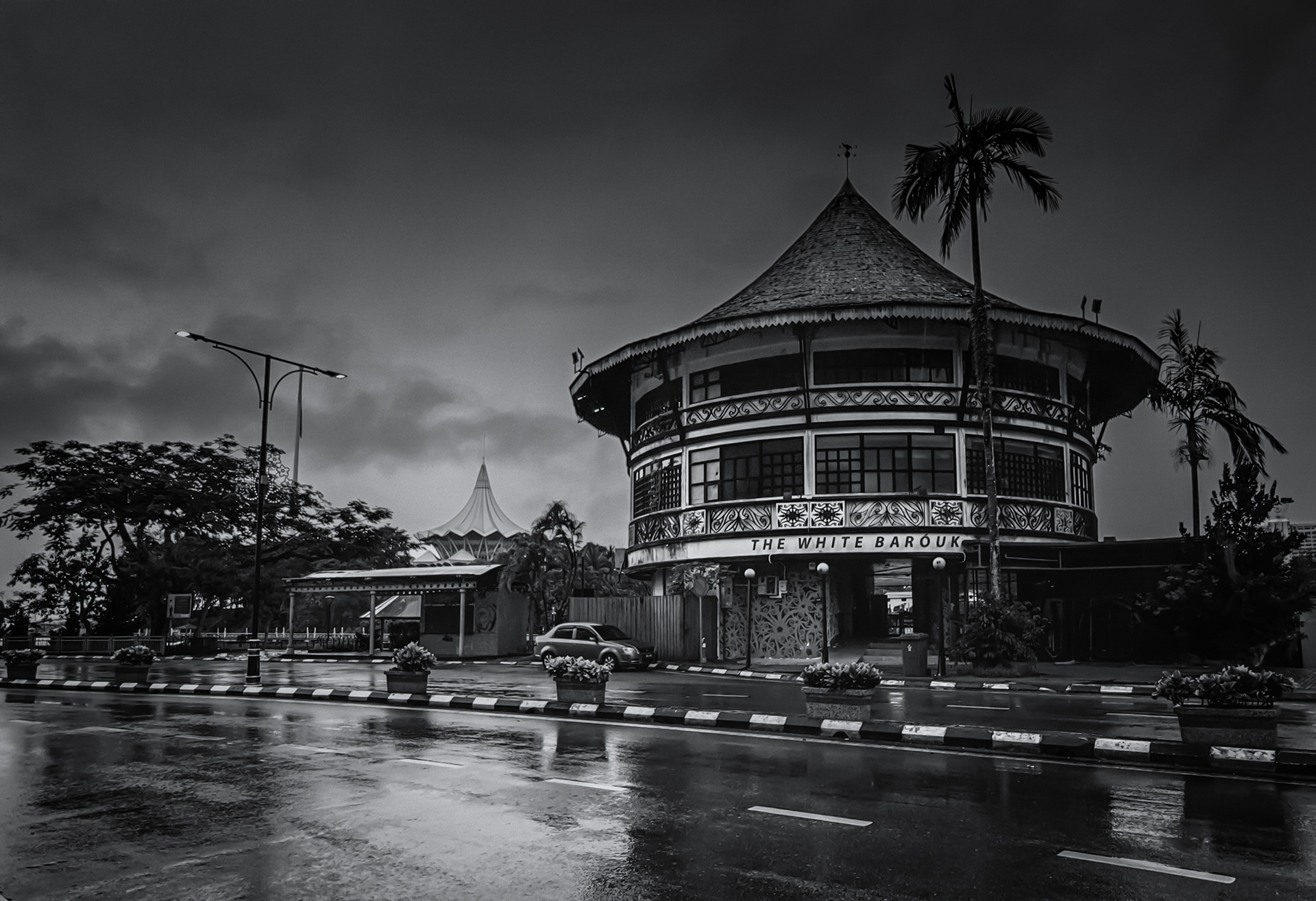 Black-and-white view of distinctive curved architecture beside a rain-soaked city street, beneath a dark and dramatic overcast sky.