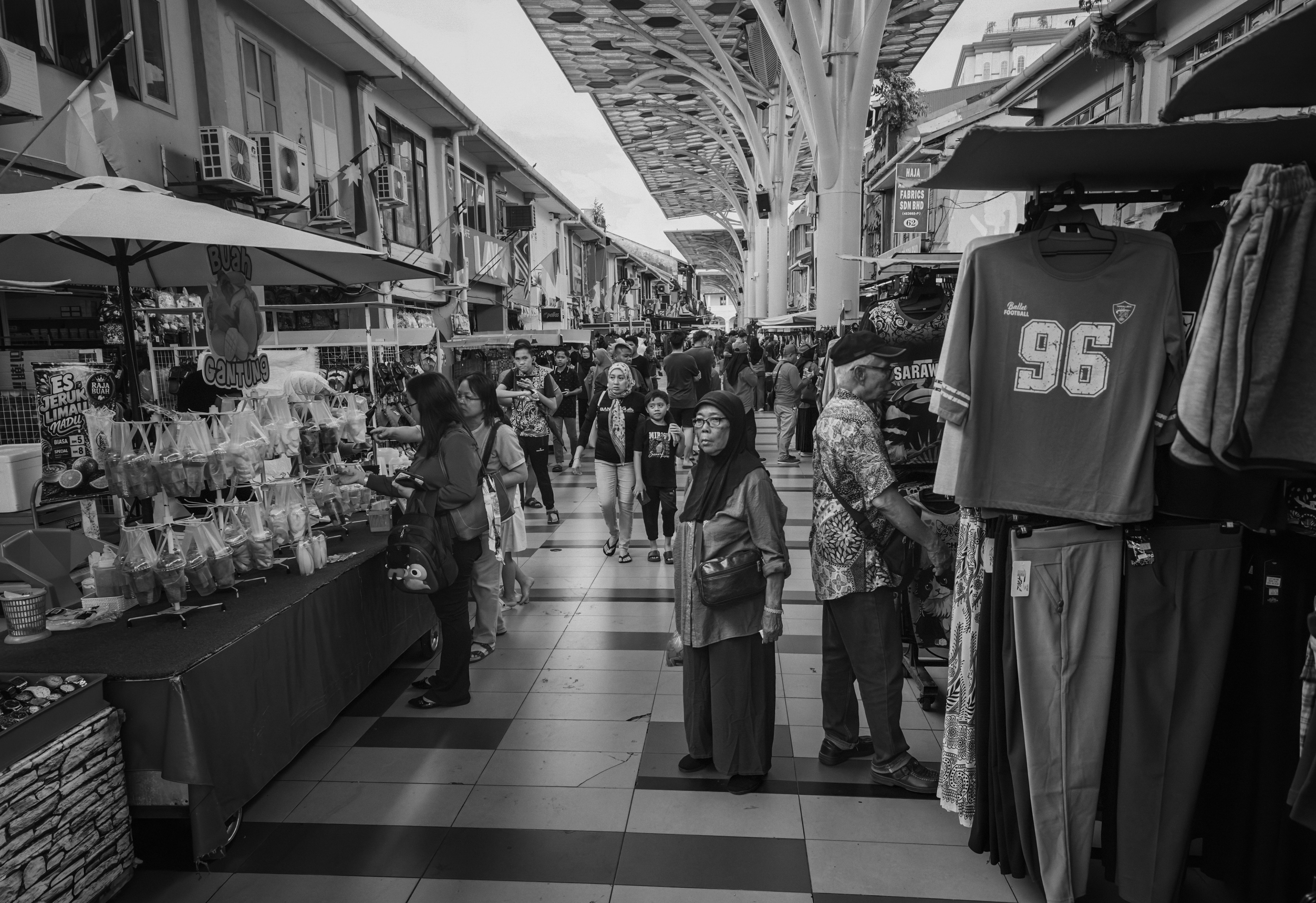 A black-and-white street photograph of a bustling covered market with shoppers browsing stalls and walking through a wide pedestrian arcade. Clothing displays, food vendors, and modern architectural canopies frame the lively scene, capturing everyday life at India Street, Kuching, Sarawak.