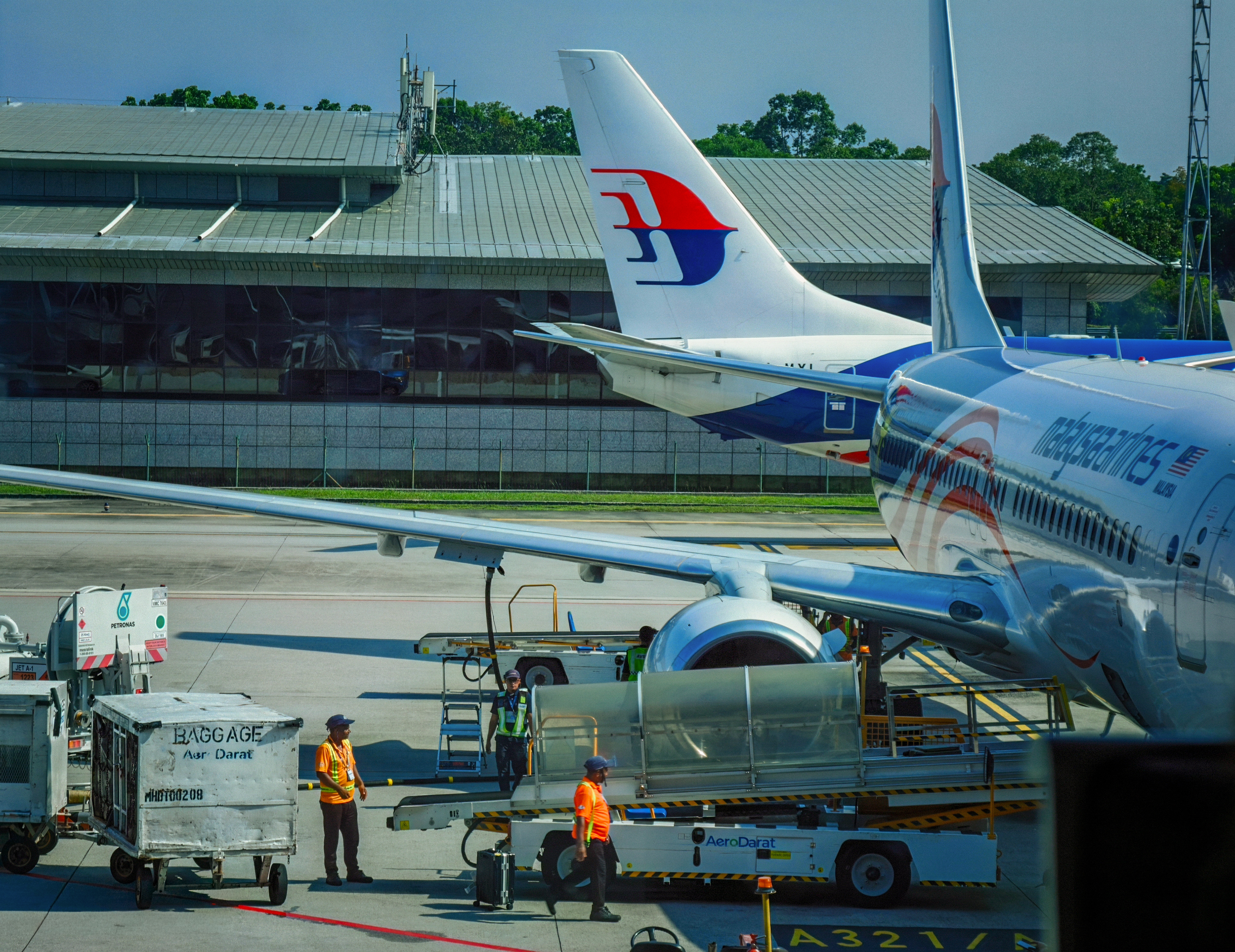 Ground crew prepare a Malaysia Airlines aircraft for departure on the airport apron, with service vehicles, baggage carts, and airport equipment surrounding the aircraft. The terminal building and another Malaysia Airlines tail fin form the backdrop, highlighting the coordinated activity at Kuala Lumpur Airport, Malaysia.