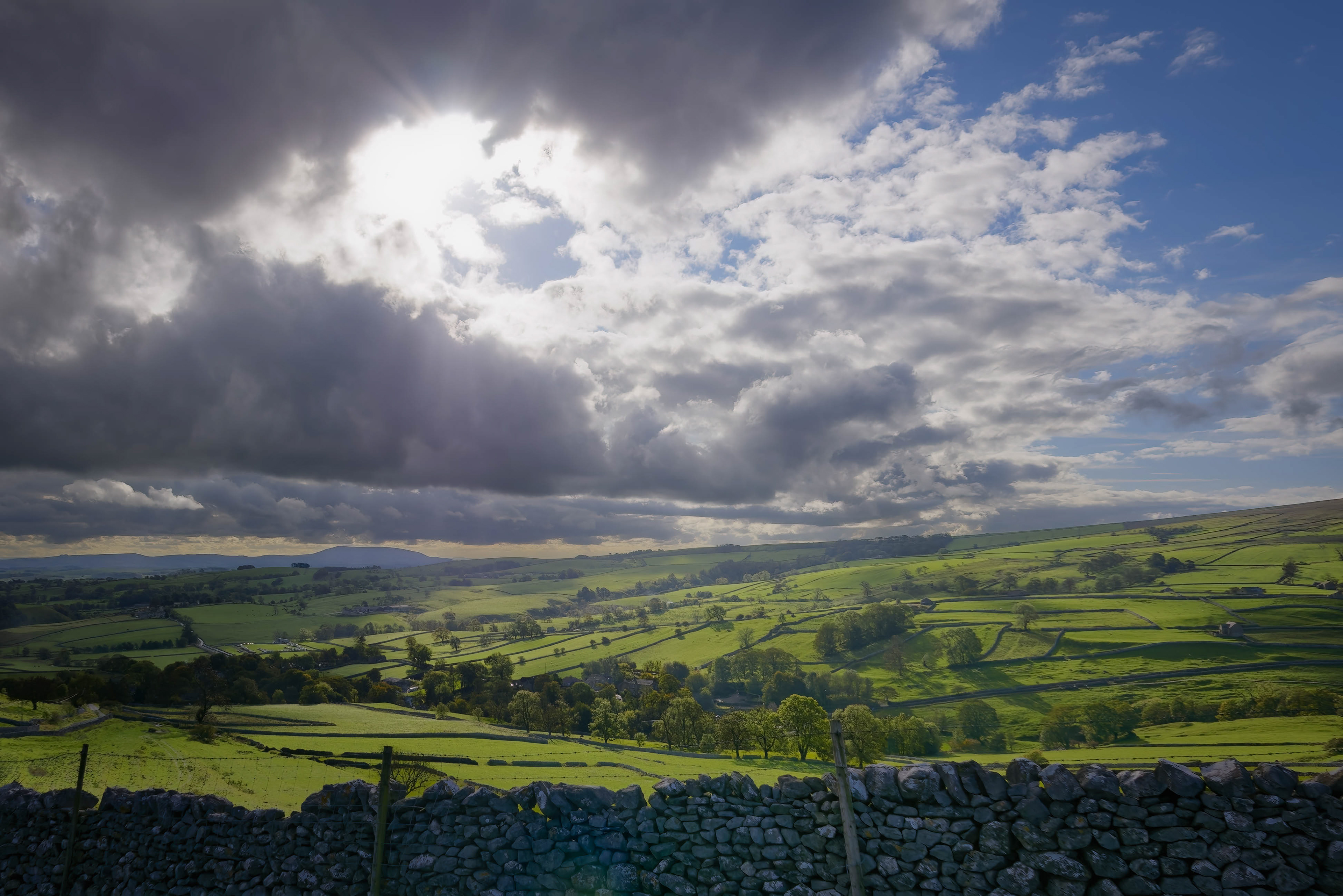 A dramatic landscape photograph of vibrant, rolling green fields divided by ancient dry stone walls, illuminated by a brilliant burst of sunlight breaking through a heavy, darkly clouded sky.