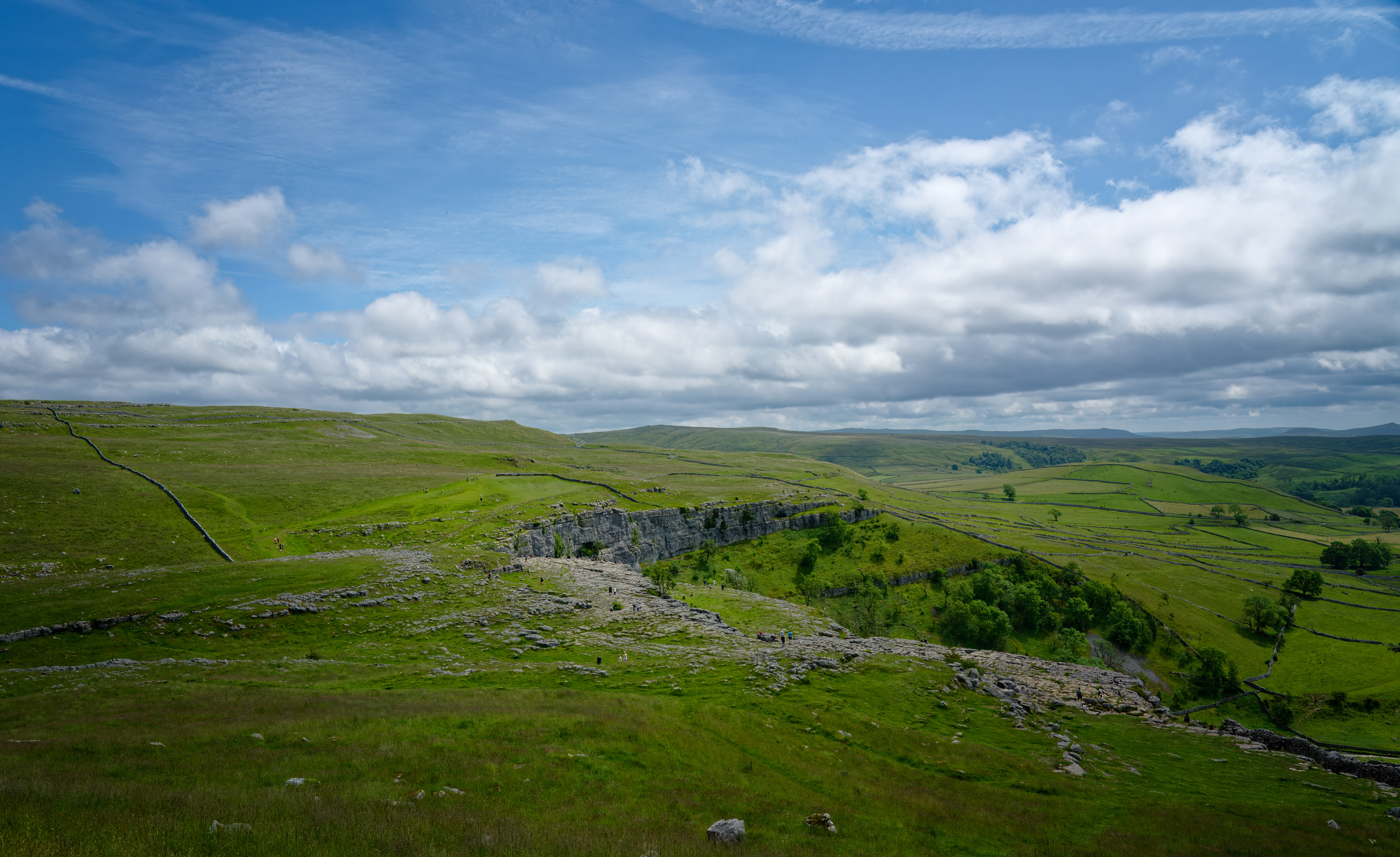 A sweeping fine-art landscape photograph showing a vast, rolling green valley featuring a prominent, cracked limestone pavement at the edge of a cliff, dotted with tiny distant hikers beneath a bright, partly cloudy sky.