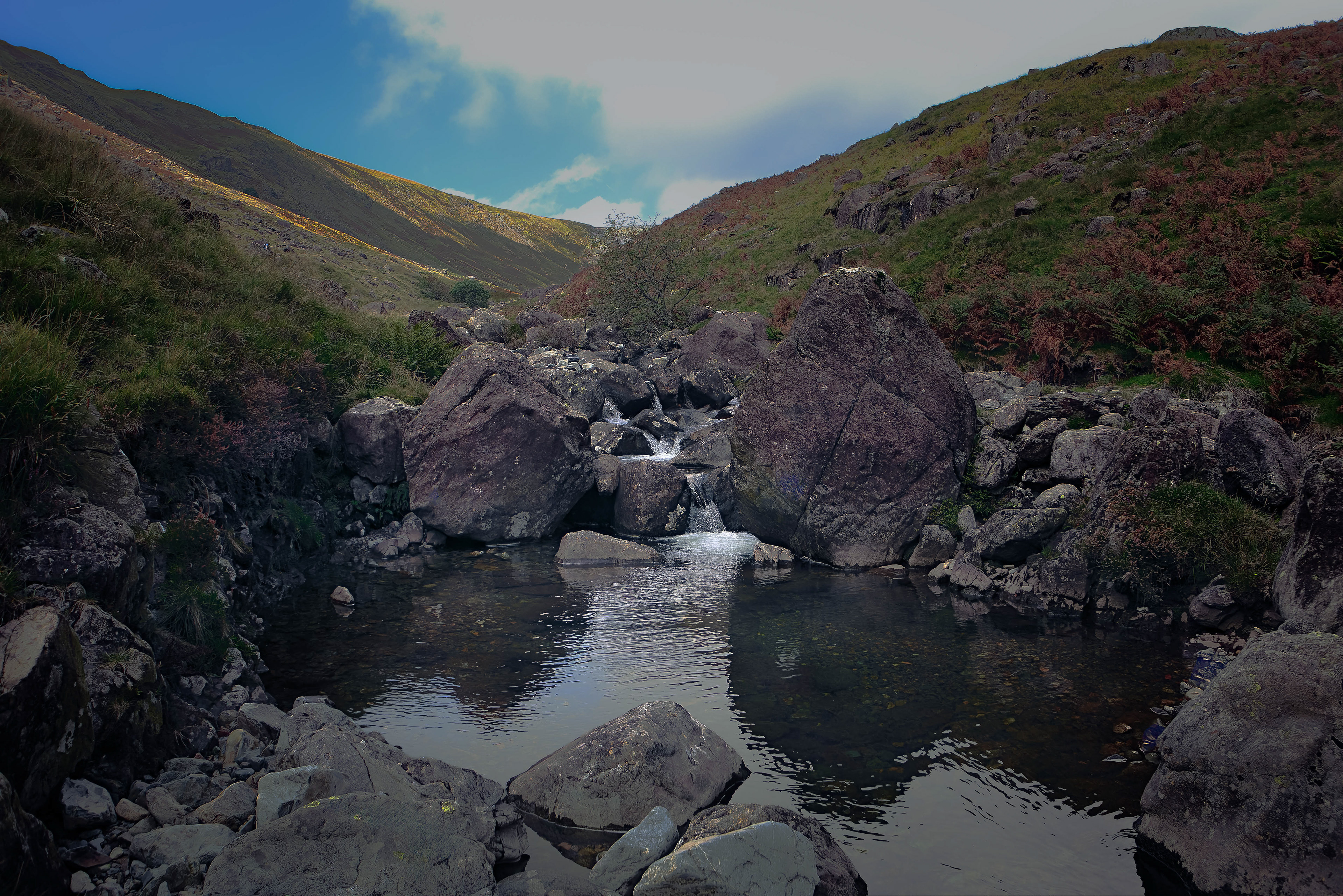 Deep still pool fed by tumbling water between great dark boulders, with sunlit green and golden fells rising in the background at Nether Beck, Wasdale.