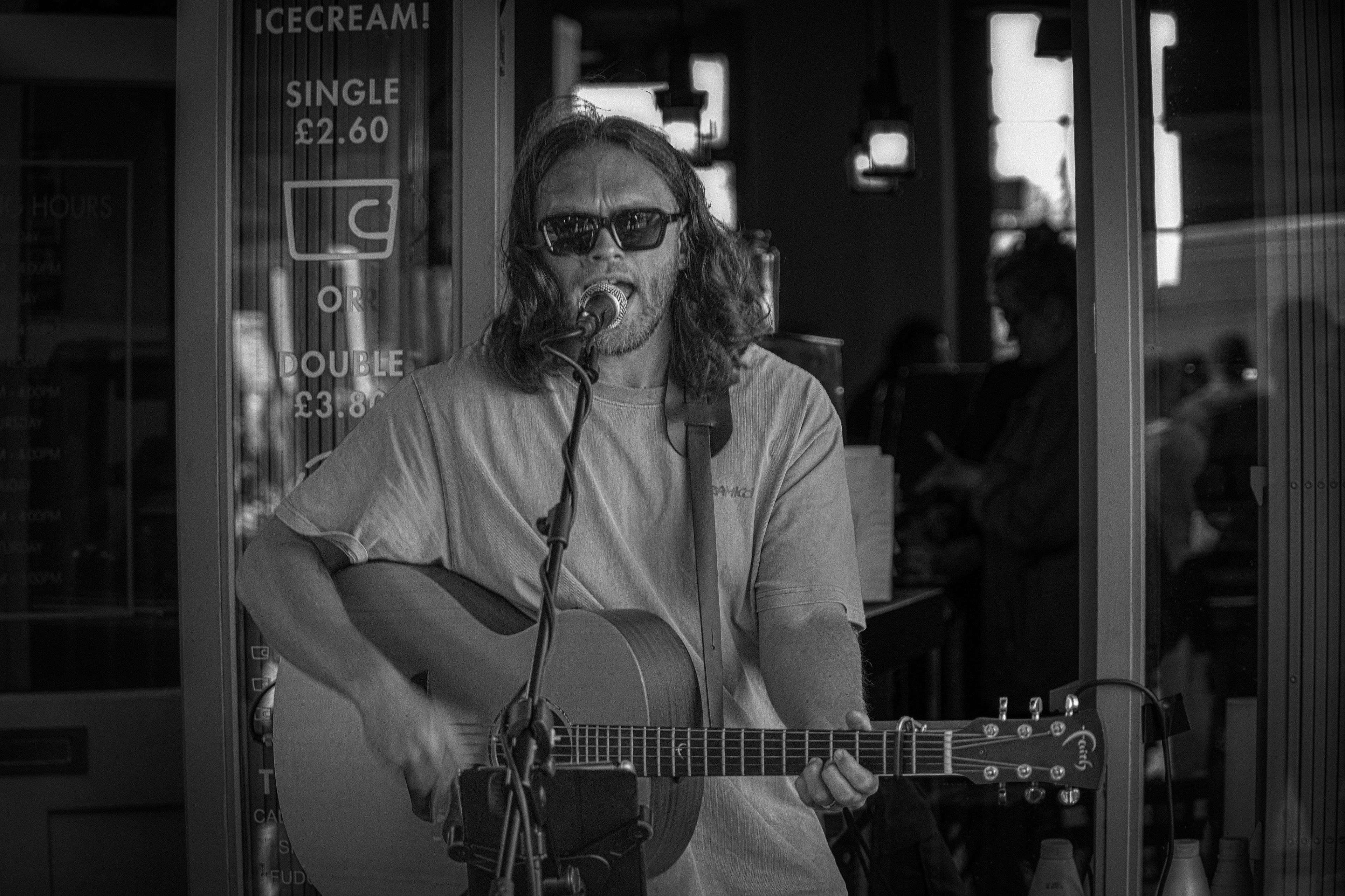 A black and white portrait of a long-haired musician wearing sunglasses, passionately singing into a microphone and playing an acoustic guitar in front of a café window displaying ice cream prices.