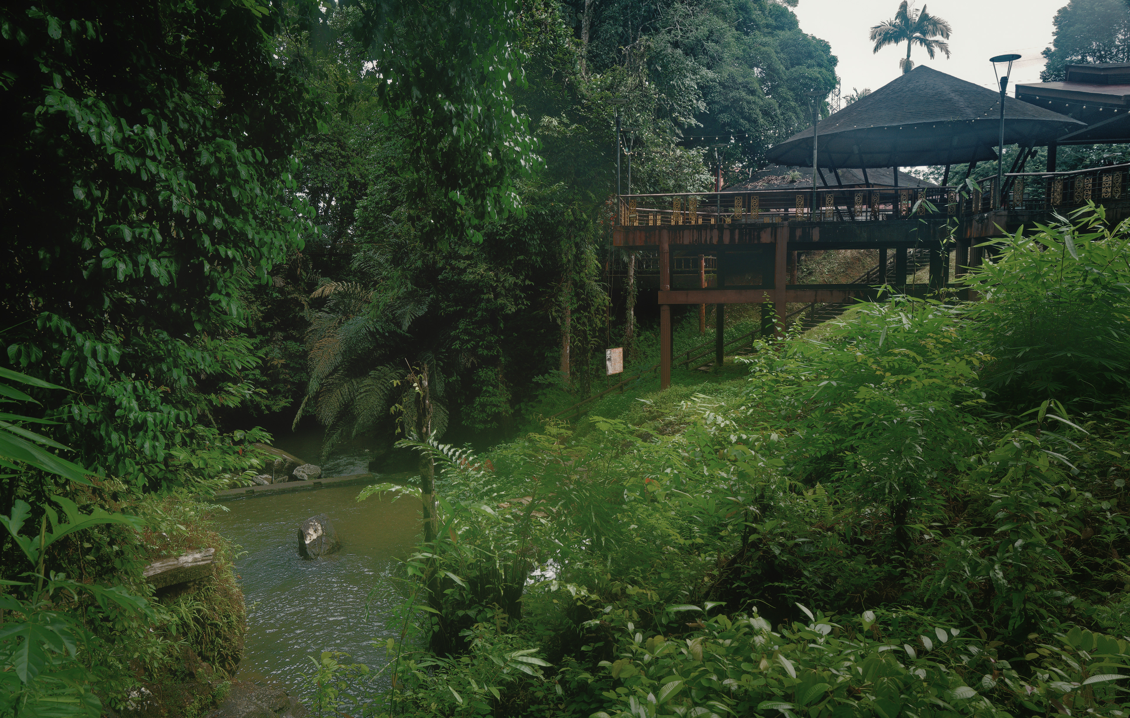 An elevated wooden pavilion overlooks a tranquil rainforest stream surrounded by dense tropical vegetation. Layers of lush greenery frame the peaceful scene, where architecture blends seamlessly into the forest landscape at Jalan Taman Rekreasi Ranchan, Serian, Sarawak.