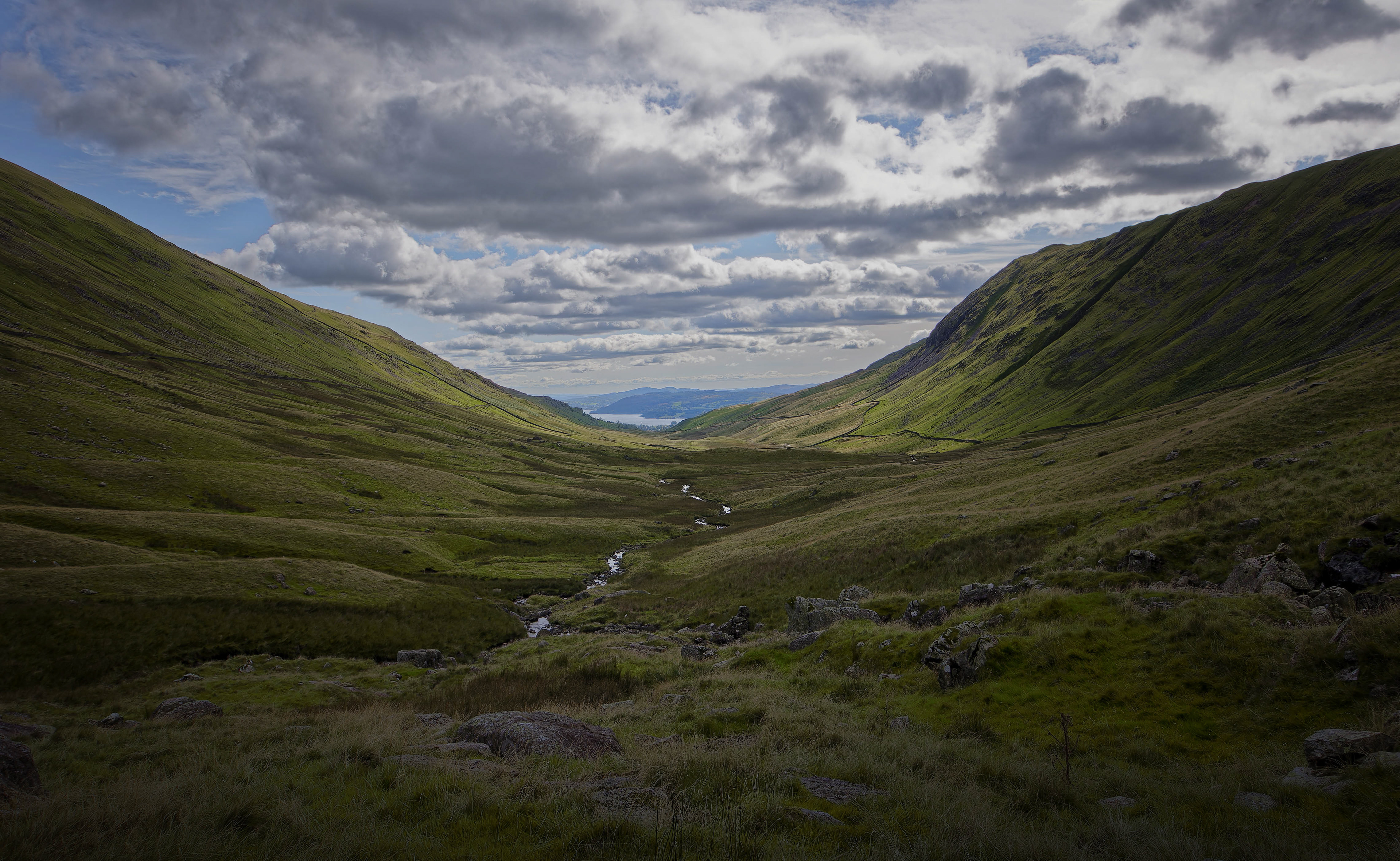 A fine-art landscape photograph looking down a vast, U-shaped green mountain valley featuring a small, winding stream in the center, stretching toward a distant body of water beneath a dramatic, heavily clouded sky casting dappled shadows across the terrain.