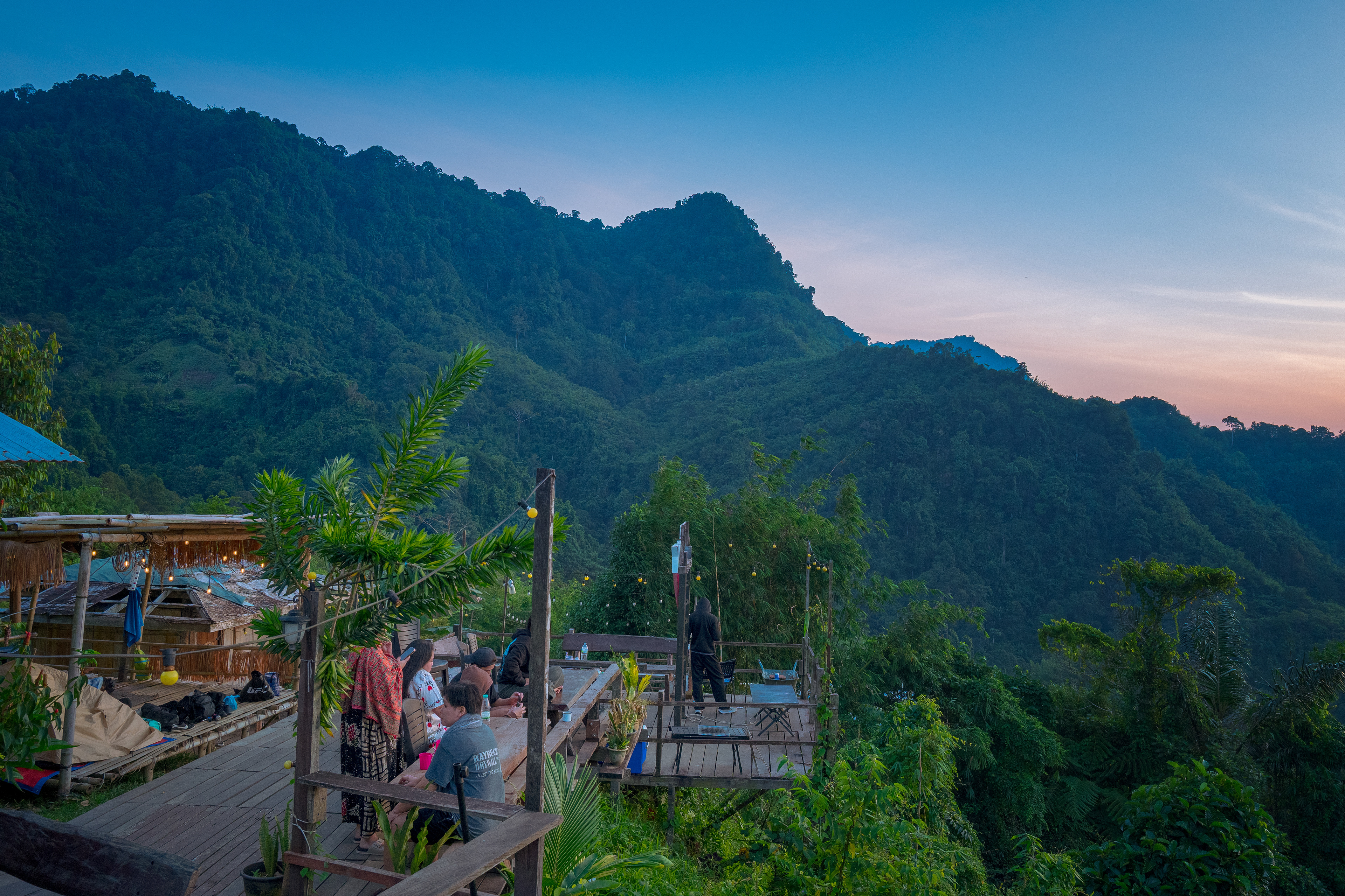 A traditional wooden stilt house with outdoor seating overlooks lush rainforest-covered mountains at sunrise. Visitors relax on elevated decks surrounded by tropical vegetation, while the soft morning sky and layered green hills create a peaceful atmosphere at the Kalimantan–Malaysia Border near Kampong Sapit, Padawan.